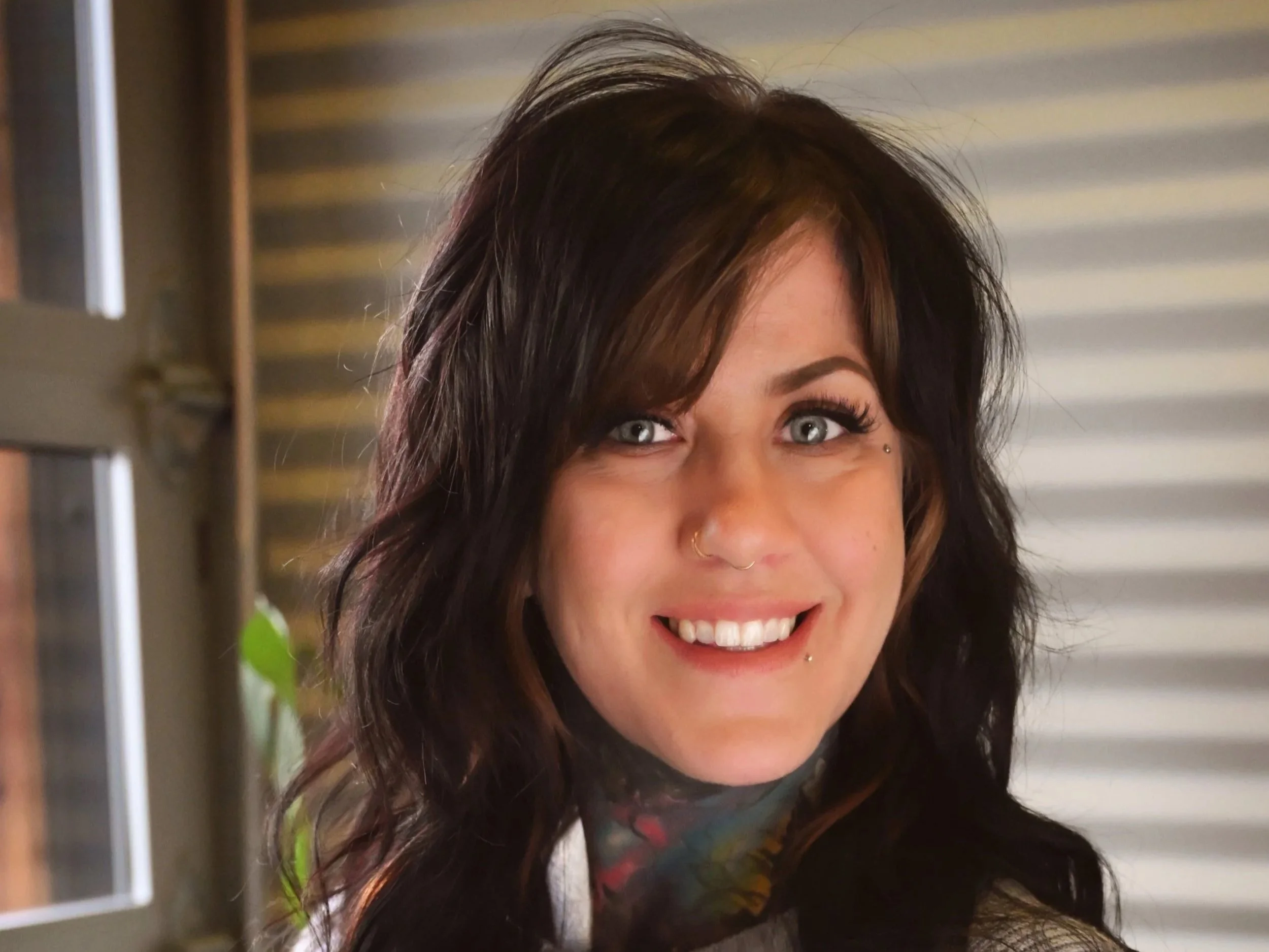 Close-up of a woman with dark wavy hair, piercing blue eyes, multiple facial piercings, and a colorful tattoo on her neck, smiling in front of a blurred background.