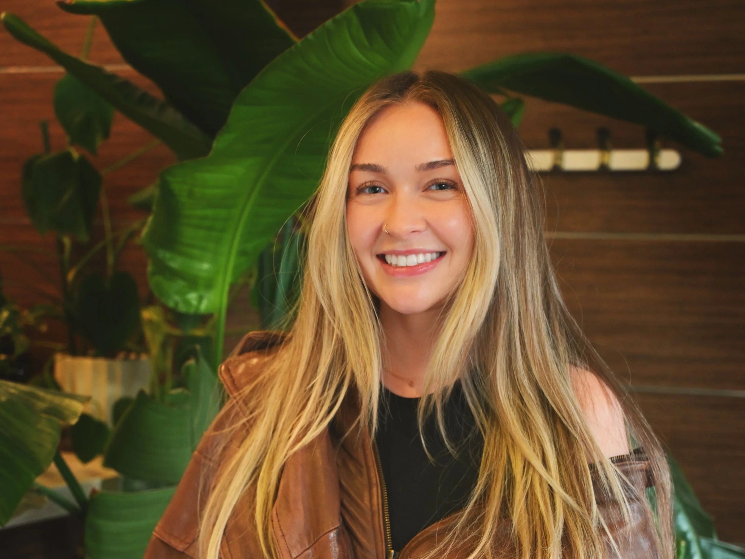 Portrait of a smiling young woman with long blonde hair, wearing a brown leather jacket, standing in front of a large green leafy plant and wooden wall.
