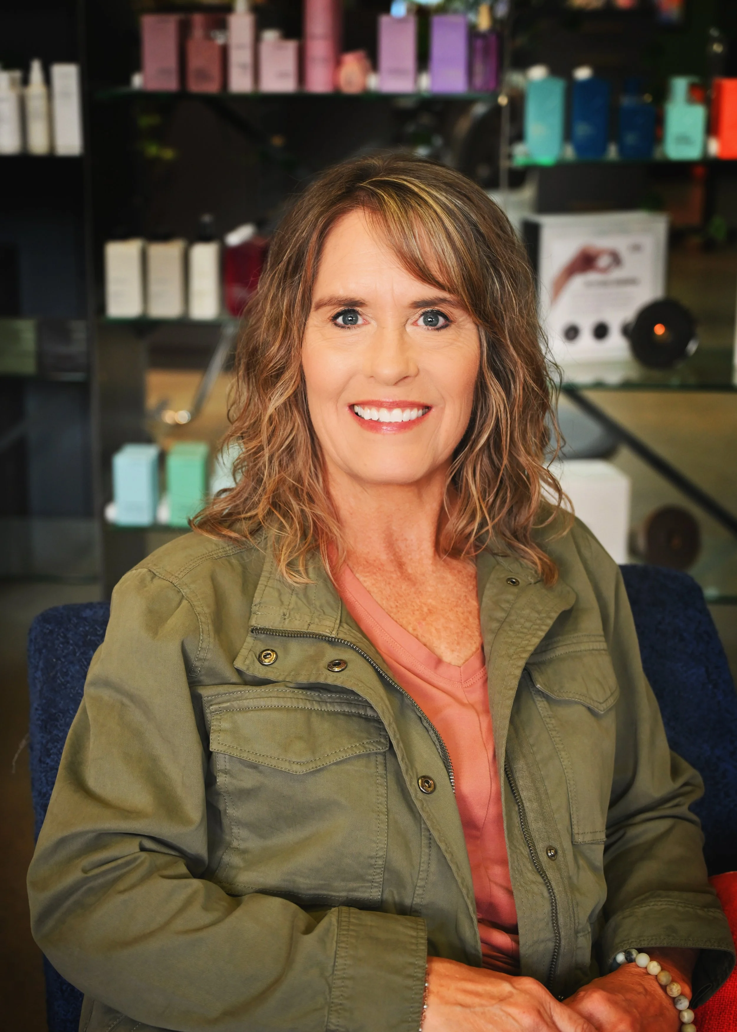 A woman with wavy brown hair and blue eyes smiling, sitting in a store or salon with shelves of hair or beauty products in the background.