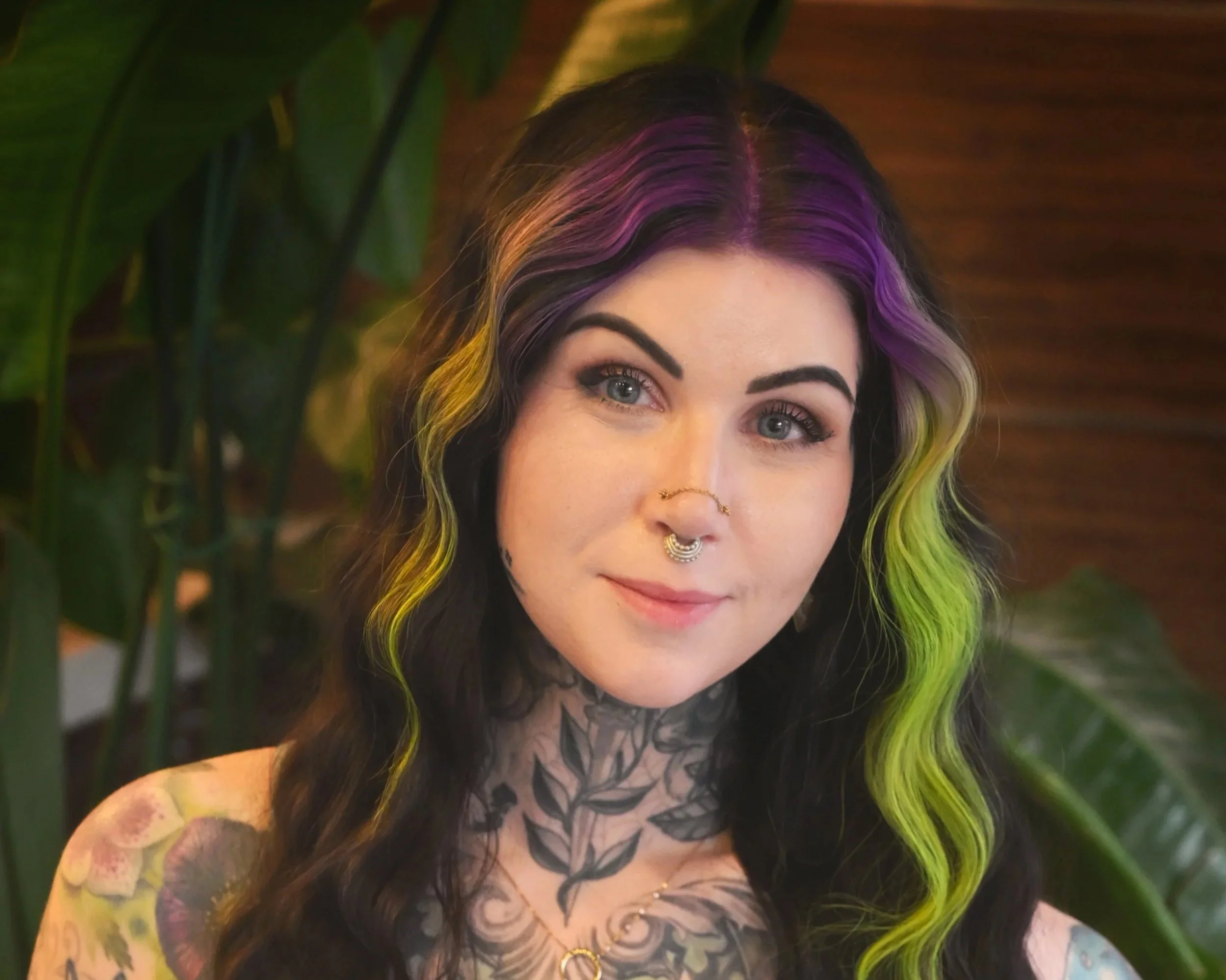 A woman with long, wavy black hair with purple and green highlights, tattoos on her shoulders and chest, and piercings on her nose and septum, smiling at the camera with a background of plant leaves and a wooden wall.