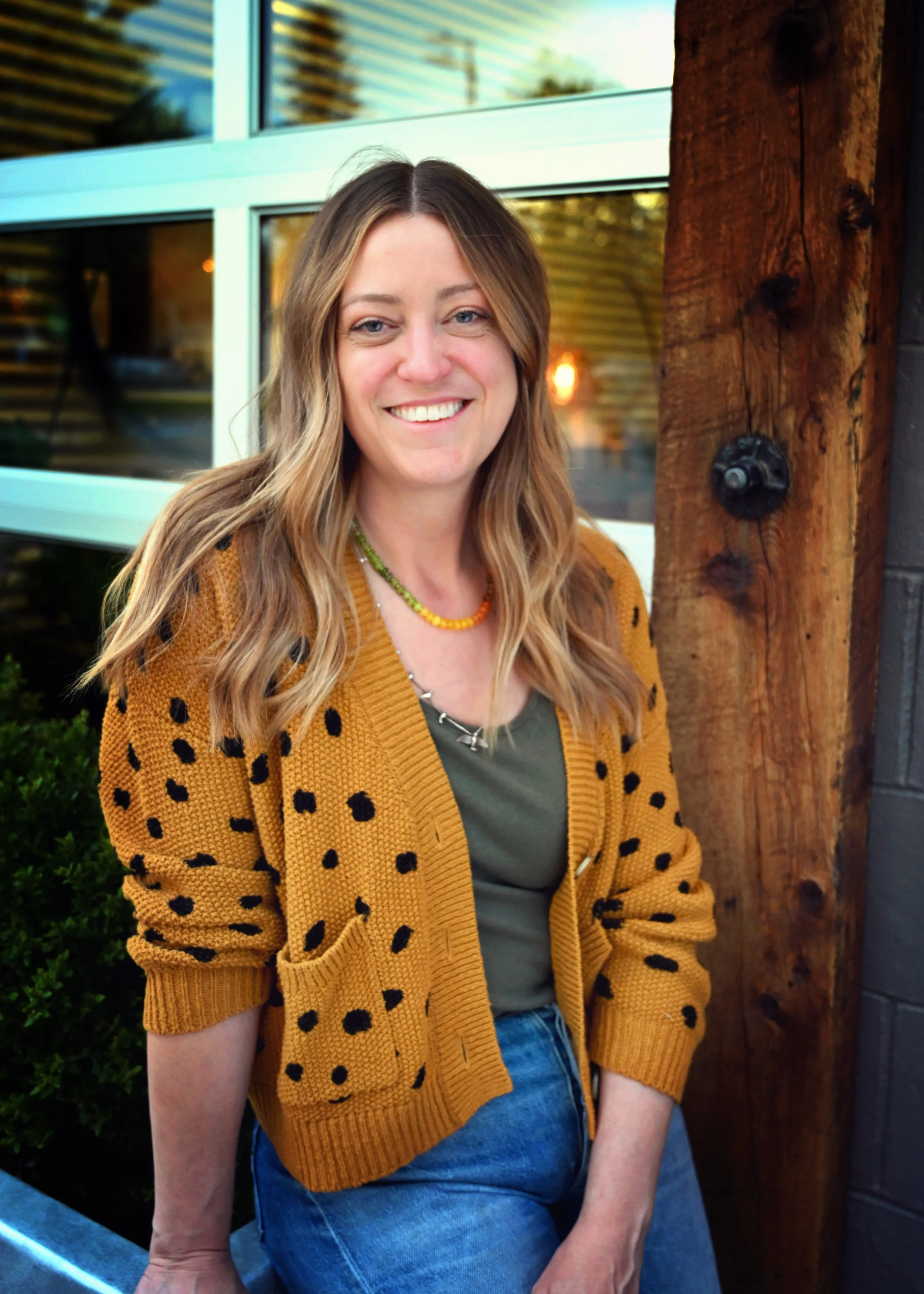 A woman with long wavy hair smiling, wearing a mustard yellow cardigan with black spots, a green top, and blue jeans, standing outside near a wooden post and a large window.