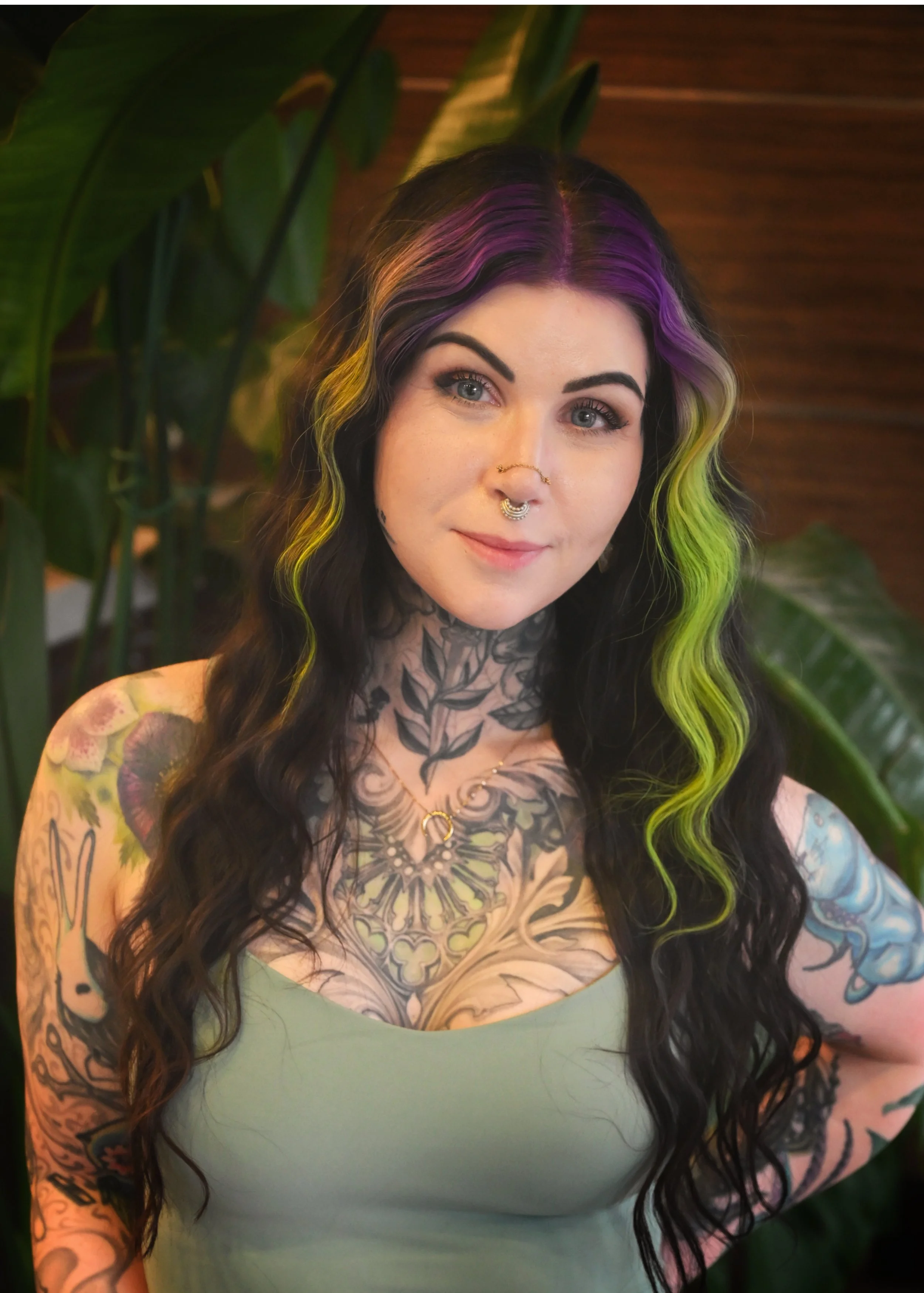 A woman with long, wavy black hair accented with purple, yellow, and green streaks, standing in front of large green leaves. She has multiple tattoos on her chest and arms, a septum piercing, and wears a light green tank top. She is smiling softly.