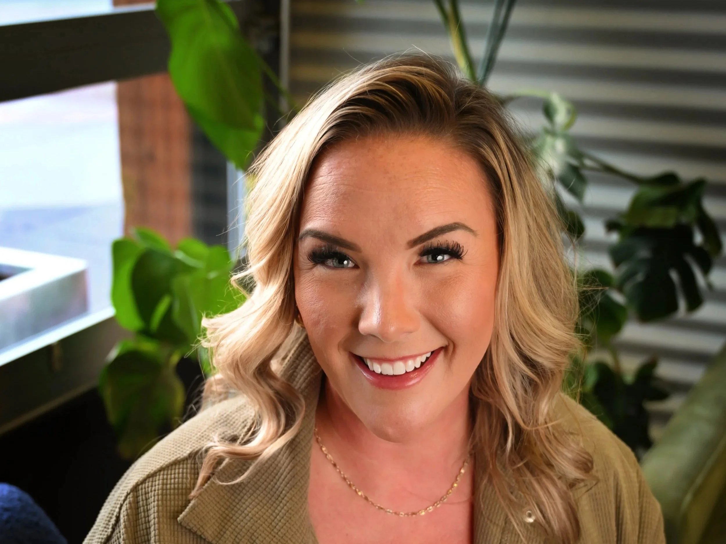Close-up of a smiling woman with blonde wavy hair, wearing a beige jacket and a gold necklace, indoors with green plants in the background.
