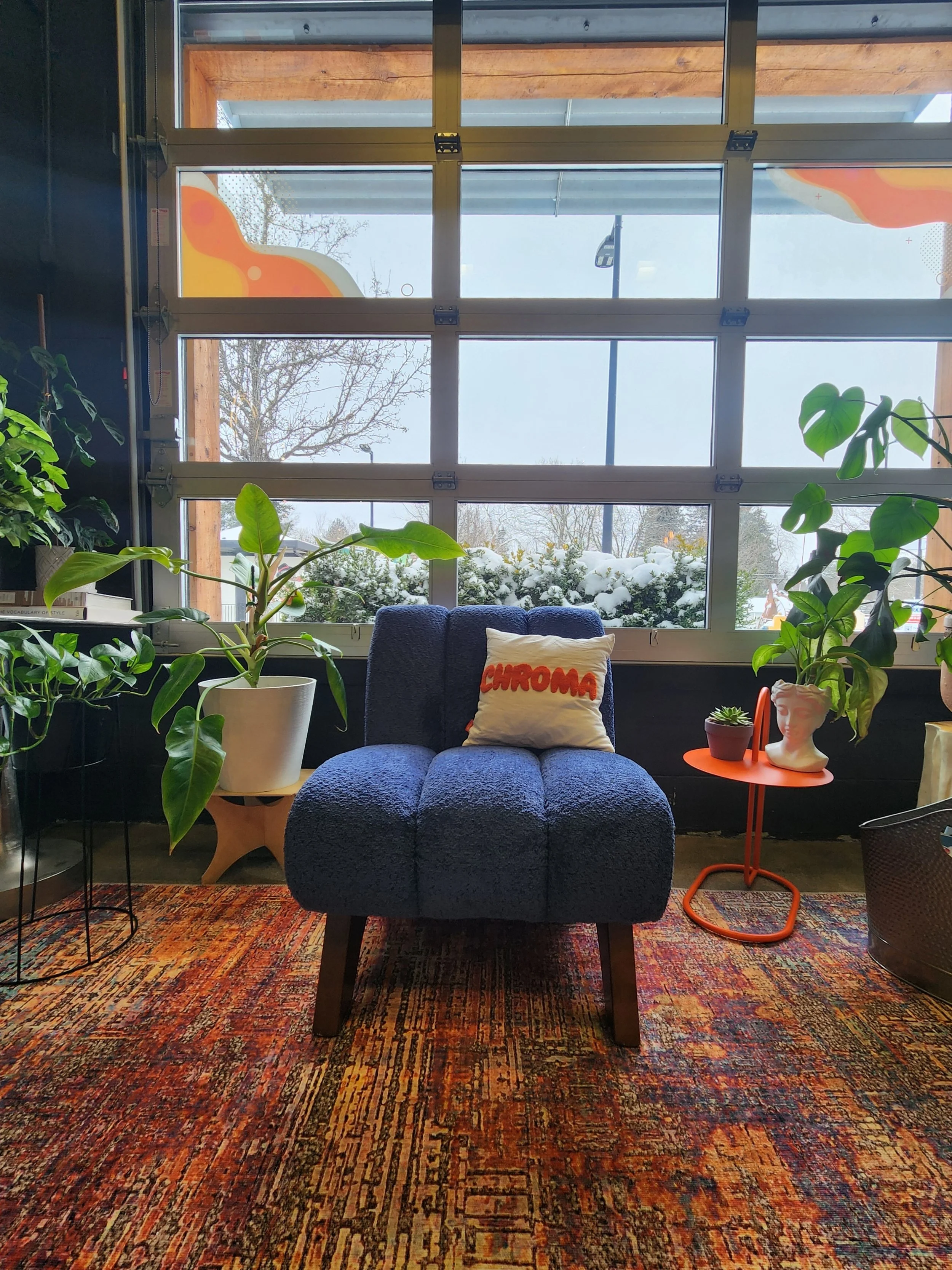 A cozy indoor sitting area with a plush blue armchair, a white decorative pillow with red writing saying 'CHROMA,' an orange side table with potted plants and a bust, surrounded by large green houseplants, and a colorful patterned rug. Large windows behind the chair reveal a snowy outdoor scene with trees.