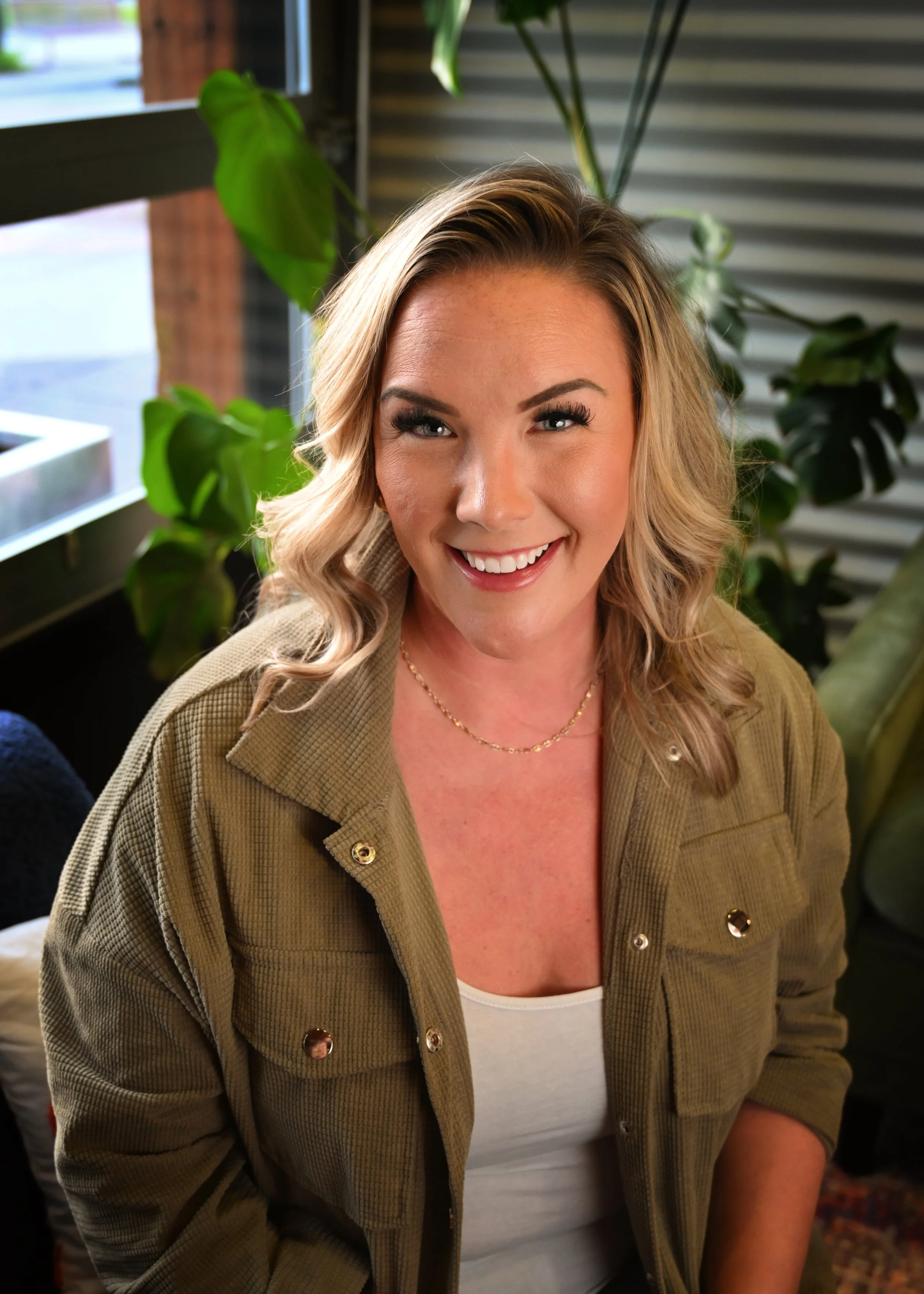 A smiling woman with blonde, shoulder-length hair styled in loose curls, wearing a beige jacket with metallic buttons over a white top, sitting indoors near a window with green plants in the background.