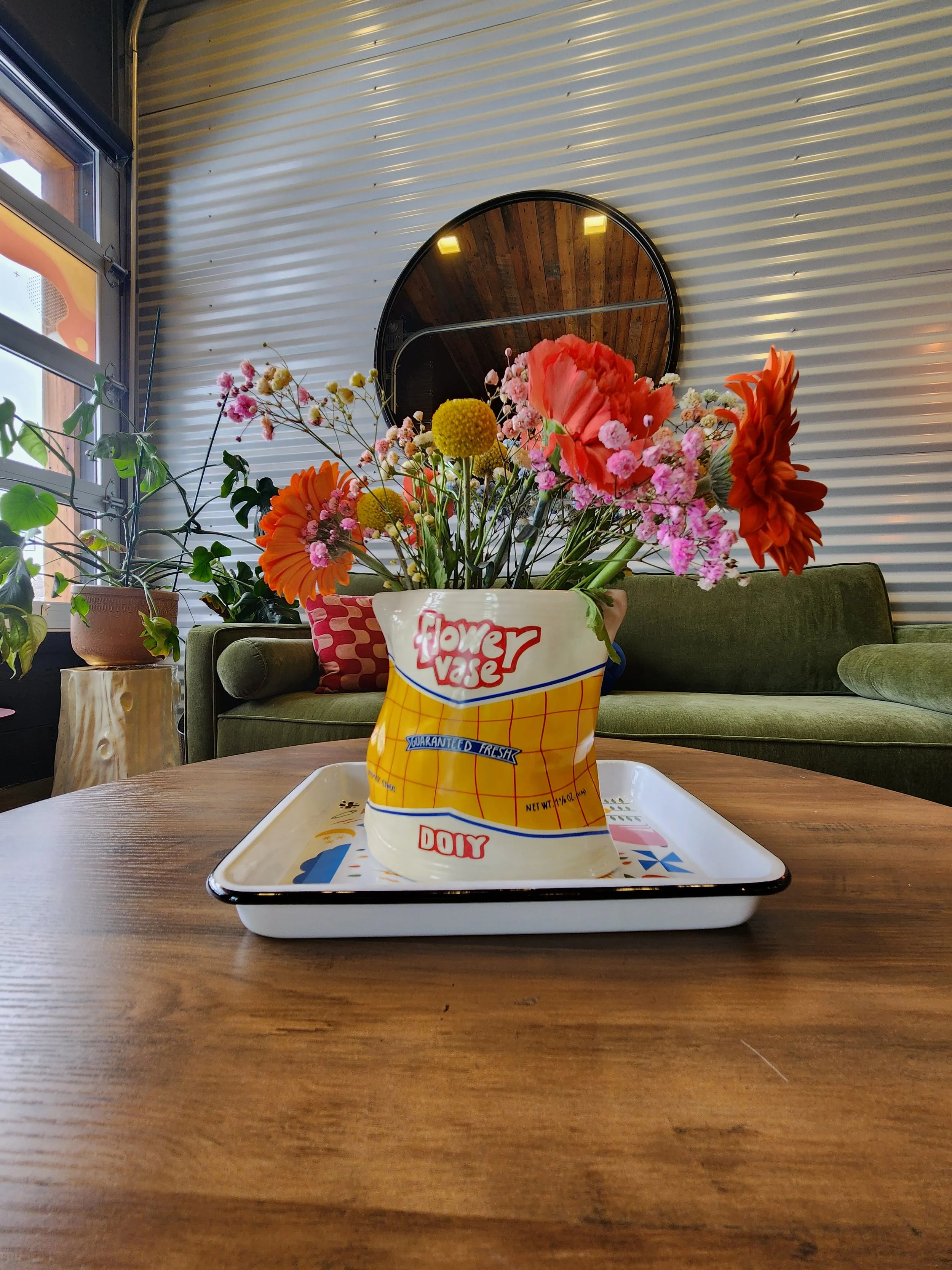 A colorful flower arrangement in a vintage-style paper bag labeled "flower vase", placed on a white tray on a wooden table in a cozy indoor setting with a green sofa and a mirror on the wall.