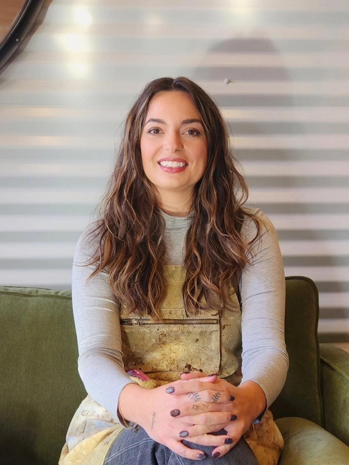 A young woman with long, wavy brown hair sitting on a green sofa, smiling at the camera, wearing a light gray long-sleeve shirt and a yellow apron.