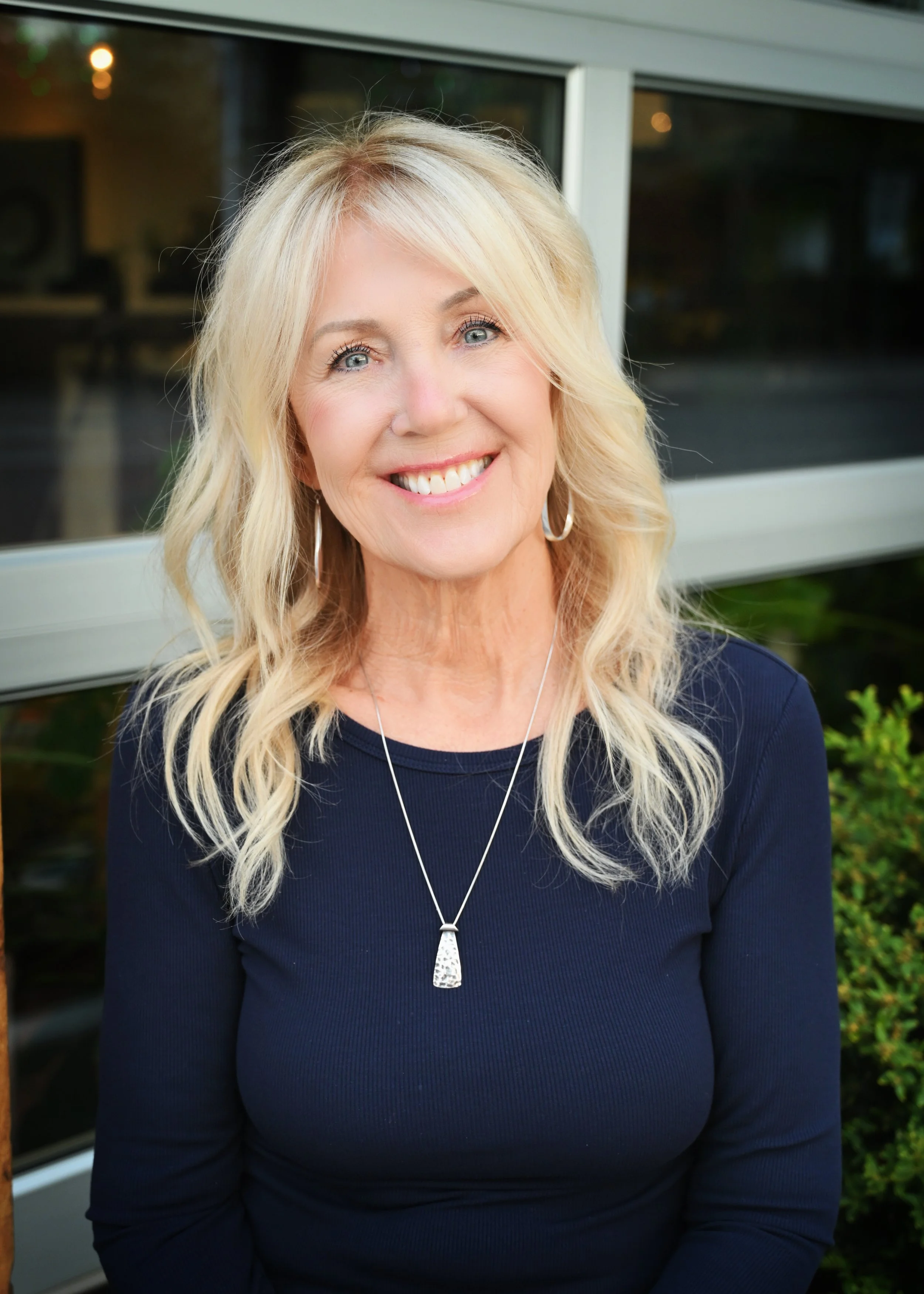 A woman with long blonde hair wearing a navy blue top, silver hoop earrings, and a silver necklace, smiling outdoors with a building and greenery in the background.
