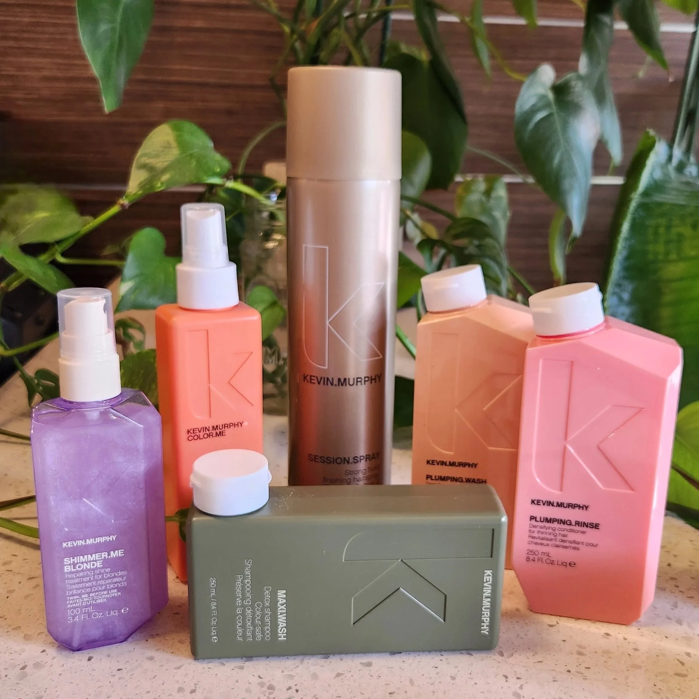 Collection of Kevin Murphy hair care products including Shimmer Me Blonde spray, Color.Me spray, Session Spray, Plumping Wash, Plumping Rinse, and Maximal Wash bar on a countertop with green leafy plants in the background.