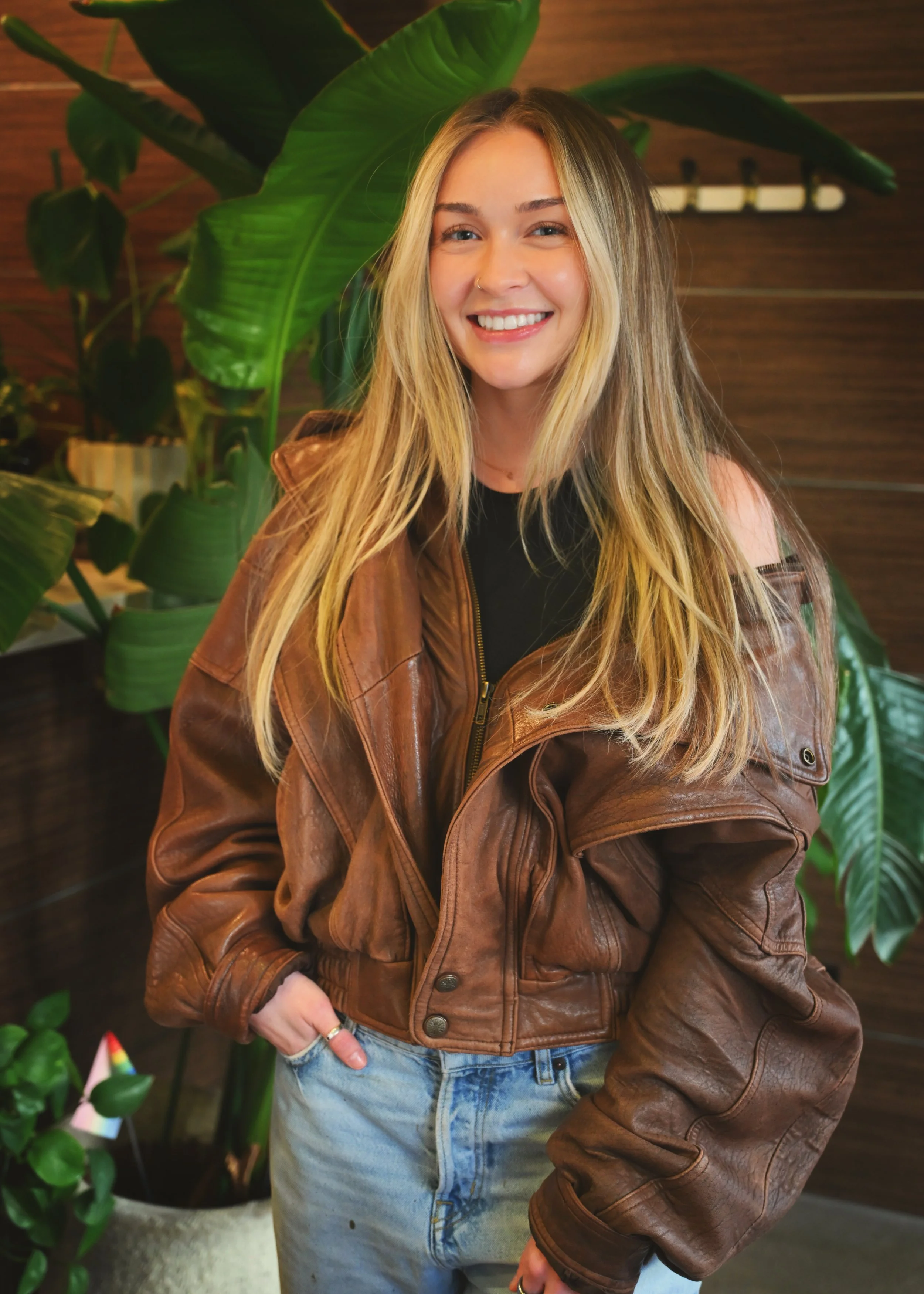A young woman with long blonde hair, smiling, wearing a brown leather jacket and jeans, standing indoors with green plants and wooden walls in the background.