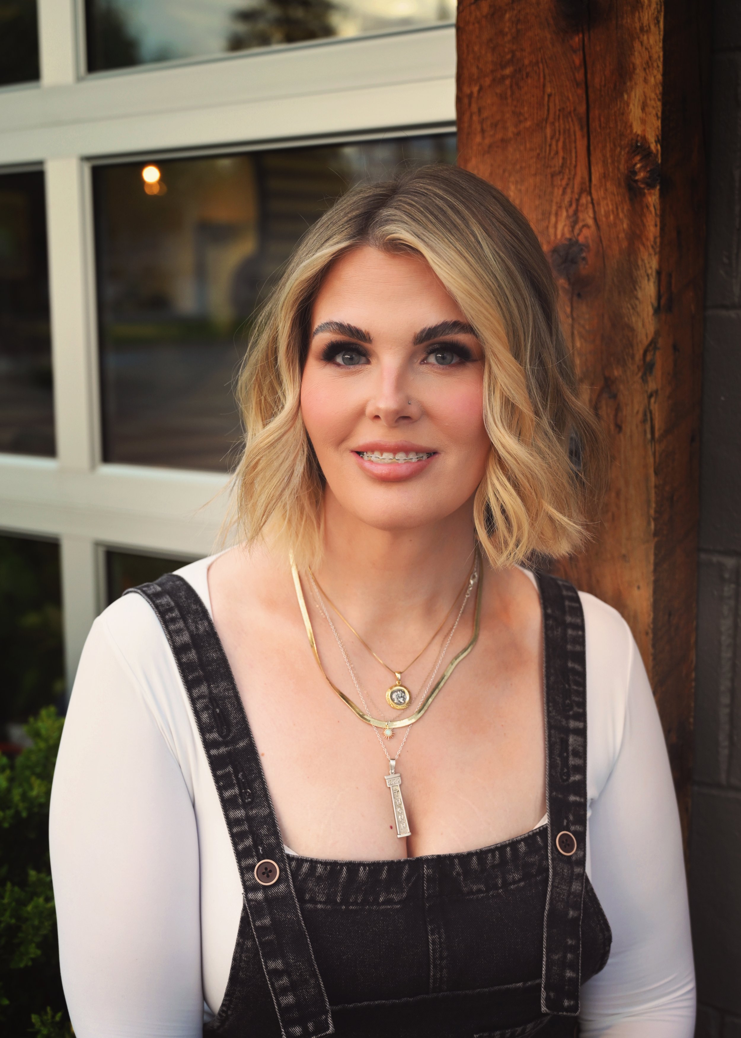 A woman with blonde hair styled in loose waves, wearing makeup, a white top with black denim overalls, and layered gold necklaces, standing outdoors near a wooden post and a window.