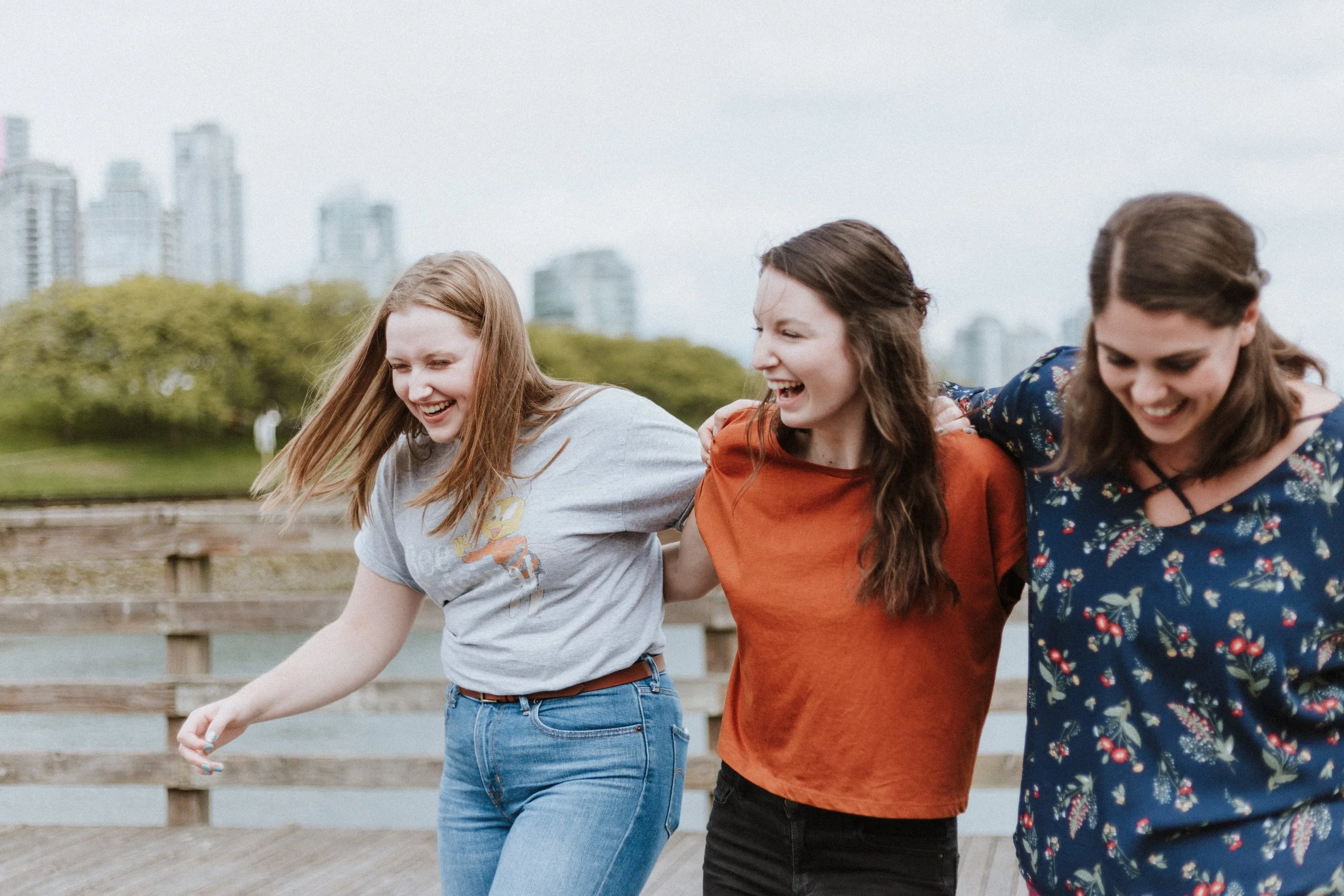  A pair of three female-presenting Caucasian young women, linking arms and laughing with a city skyline in the back. The person on the left is wearing a grey shirt with a cartoon bee, the person in the center has an orange blouse, and the person on t