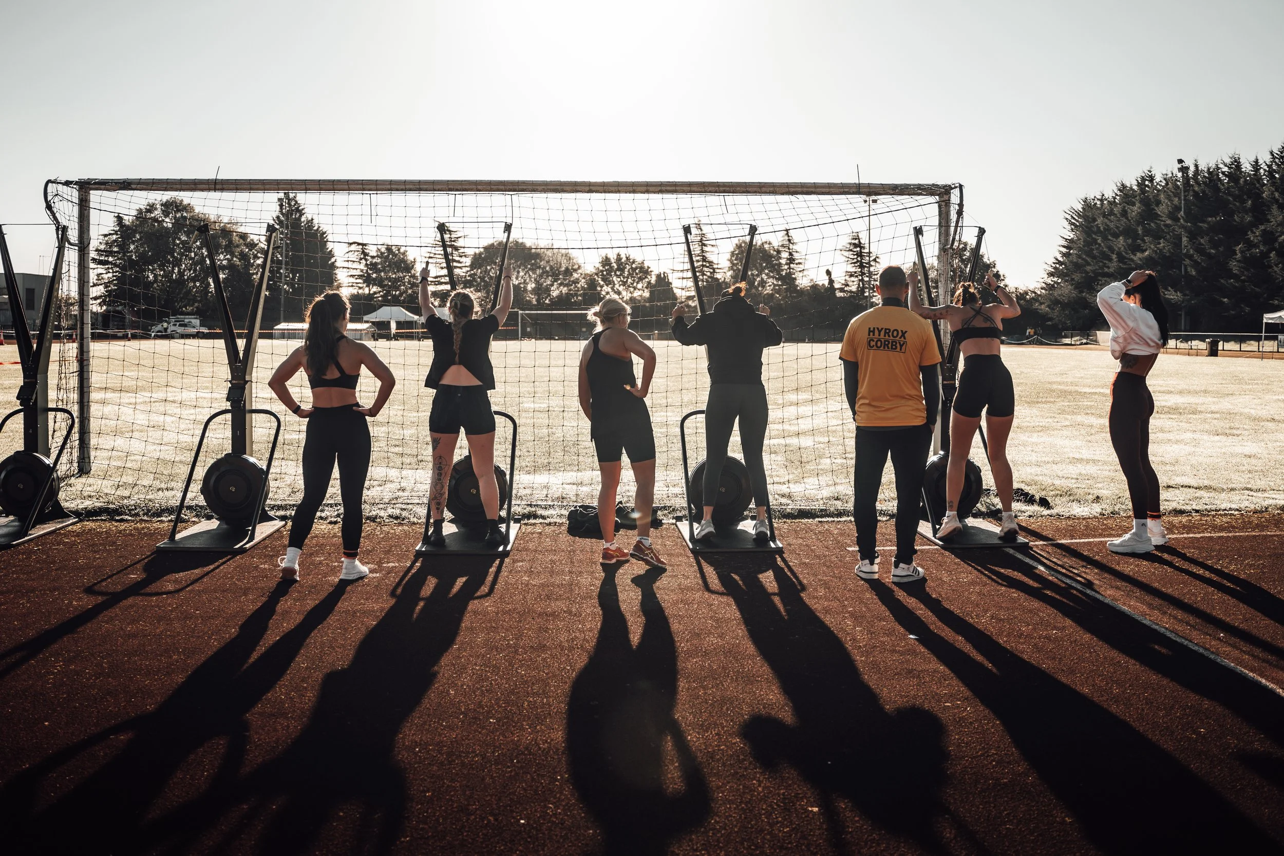 Group of people on track near soccer goal, some using workout equipment, during sunset or early morning.
