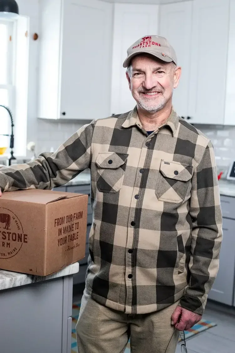 A man wearing a plaid shirt stands in front of a large cardboard box, looking directly at the camera.
