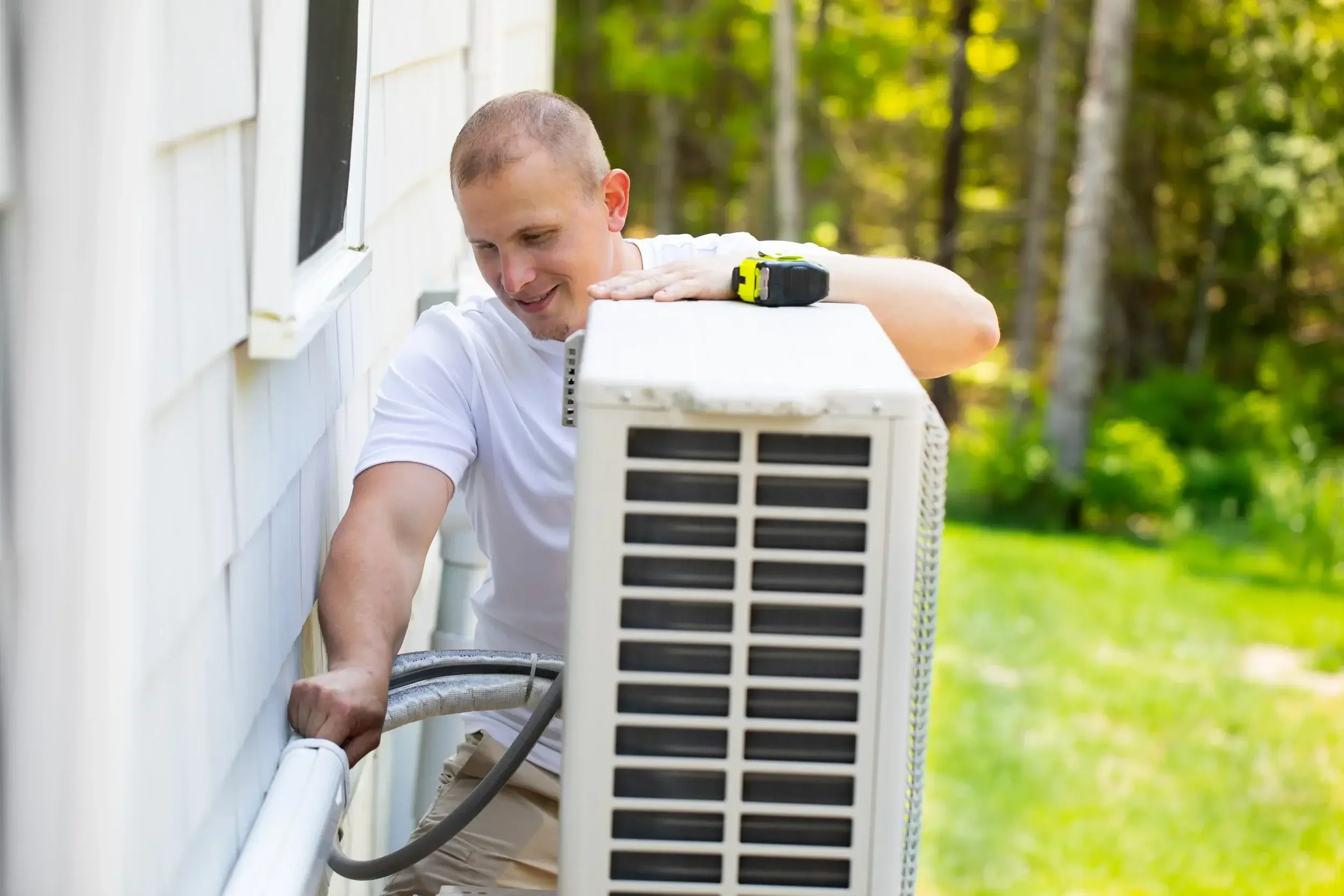 A man repairs an air conditioner unit outside, using tools and focused on his task in a residential setting.