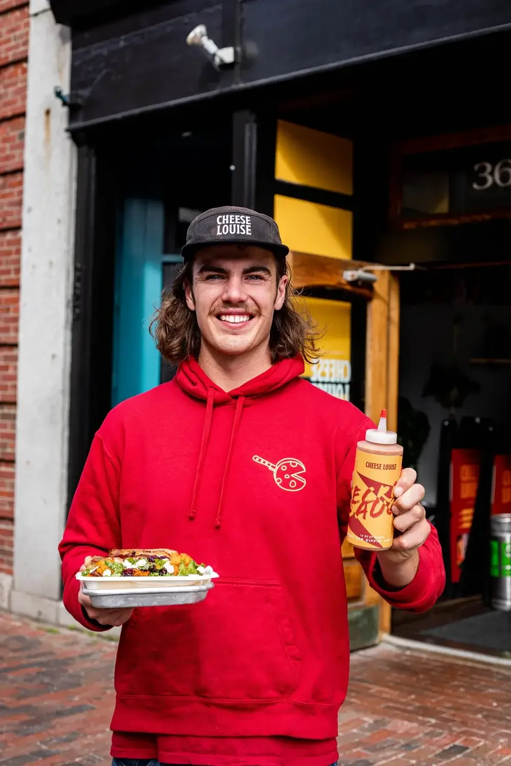  A man in a red hoodie holds a tray of food, smiling as he prepares to serve it.
