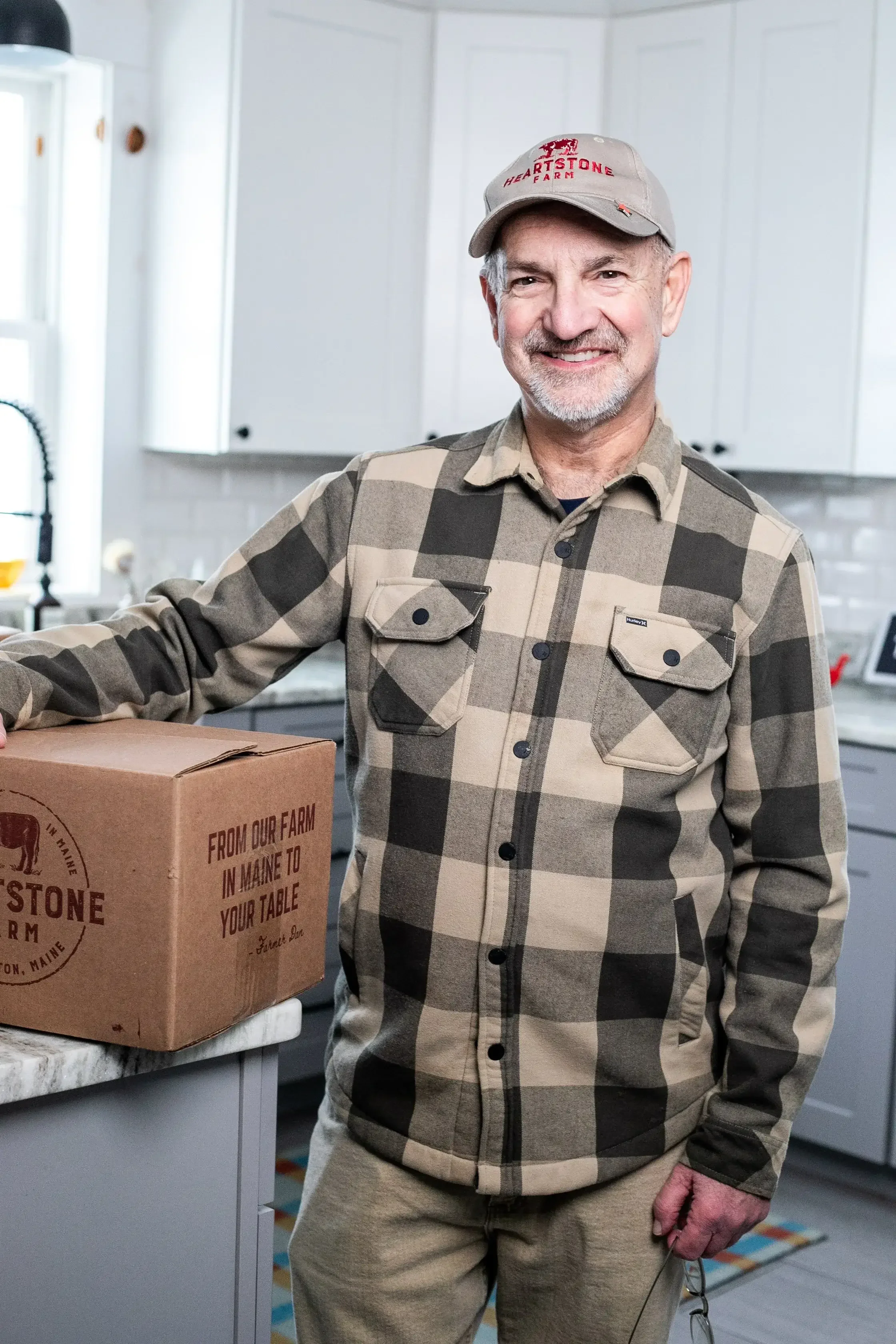 A man wearing a plaid shirt stands beside a cardboard box, looking towards the camera with a neutral expression.