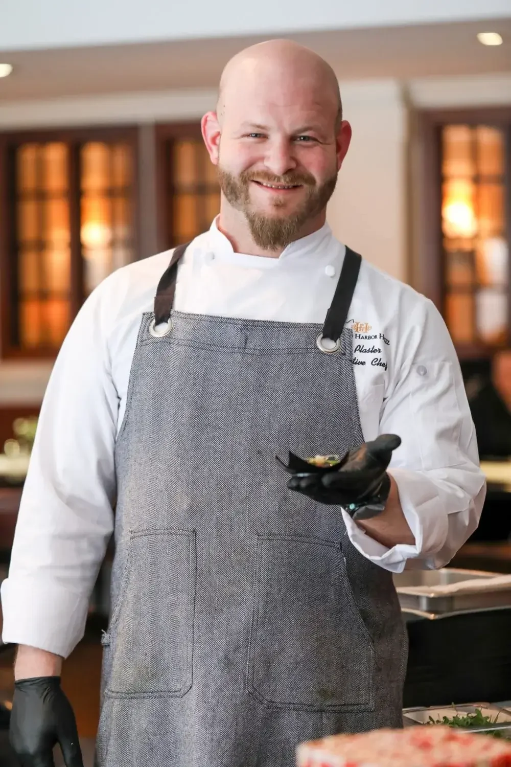 A bald man with a beard, wearing an apron, holds a piece of food, smiling in a kitchen setting.
