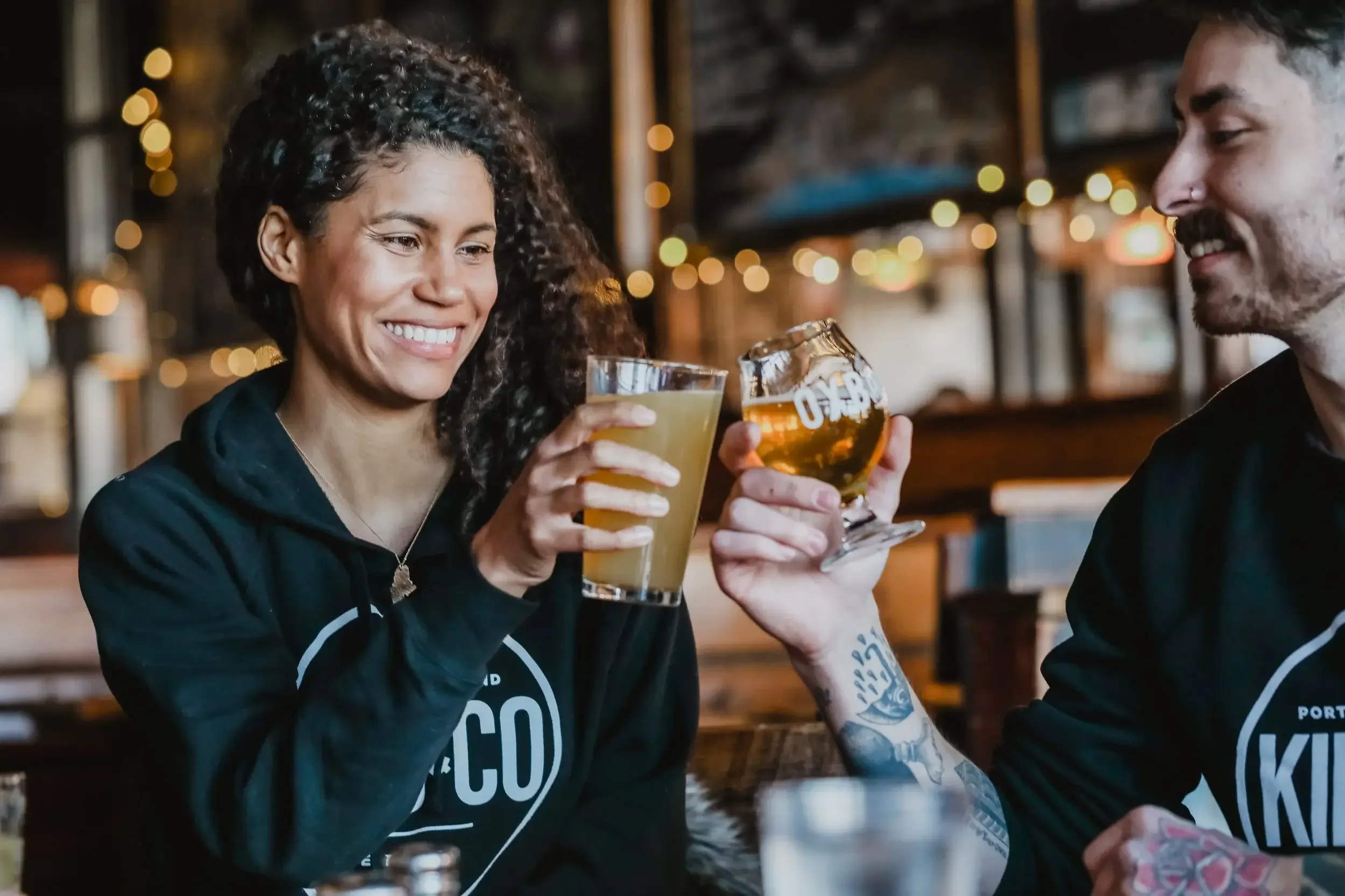 A man and woman clink beer glasses together in a celebratory toast at a restaurant table.