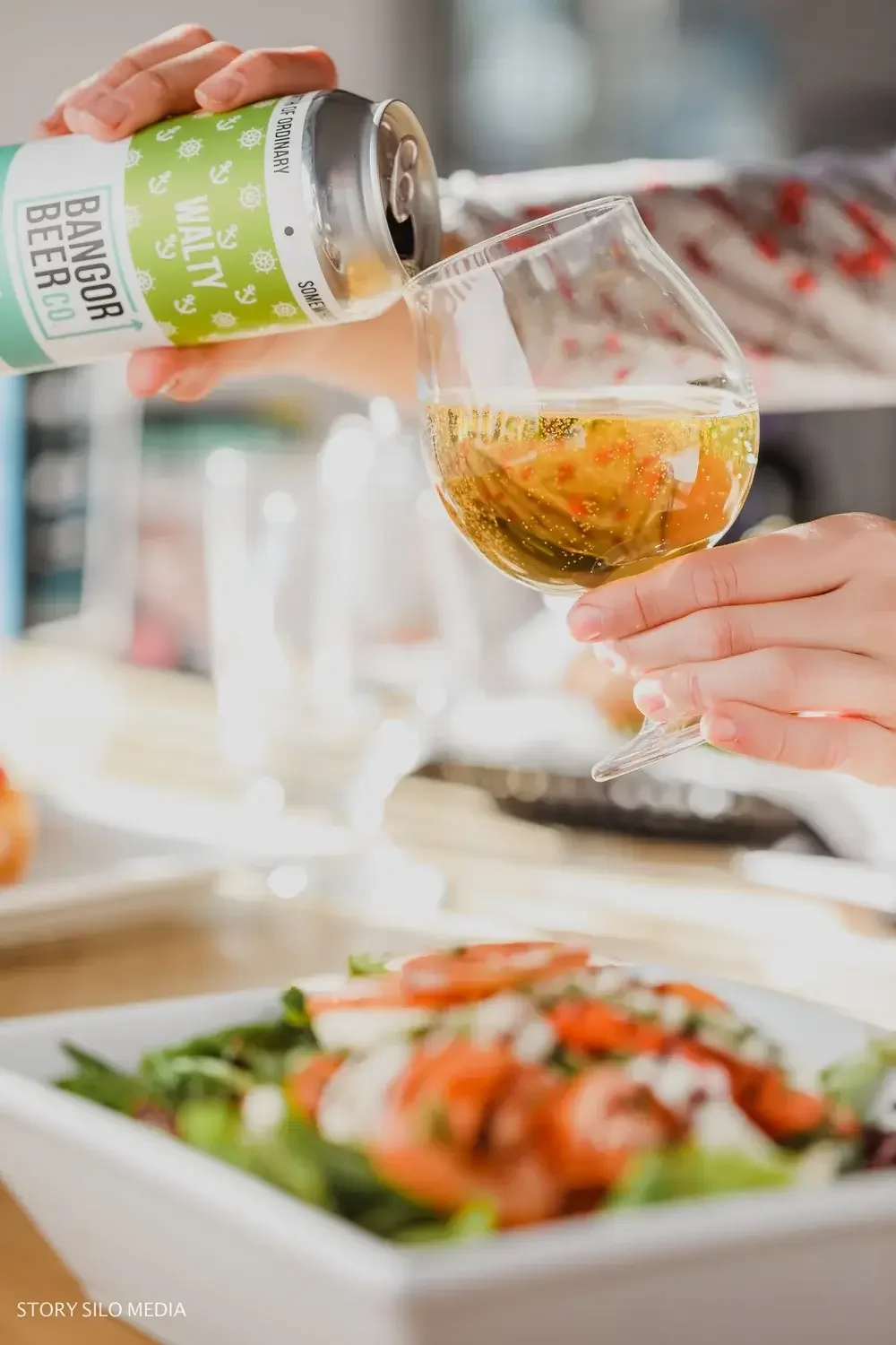 A person pours red wine from a bottle into a glass beside a fresh salad on a wooden table.