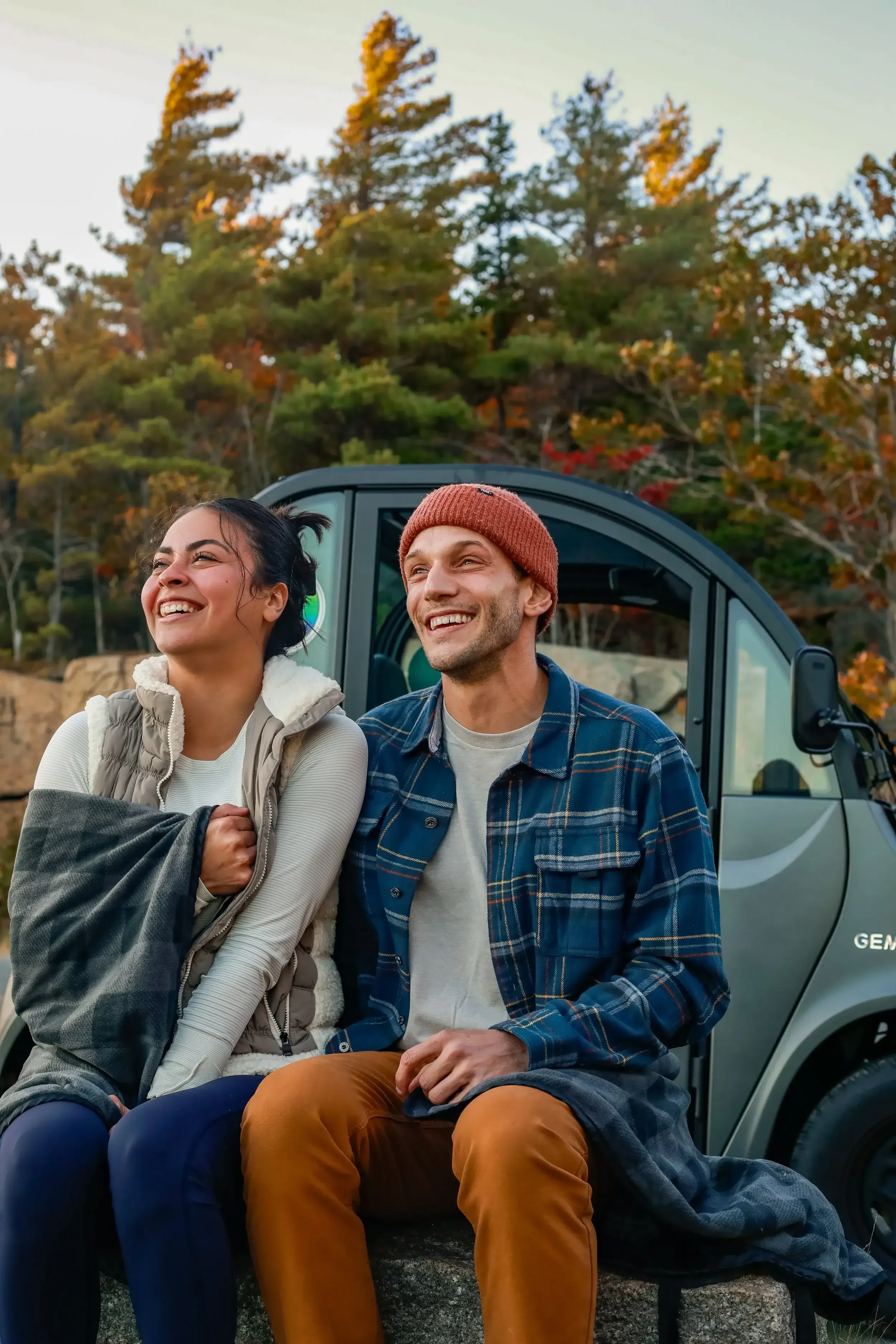 A couple sits on the back of a truck, wrapped in a blanket, enjoying a scenic outdoor view together.