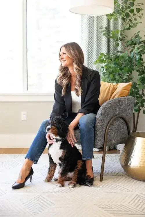 A woman sits on a chair, smiling, while her dog rests beside her on the floor.
