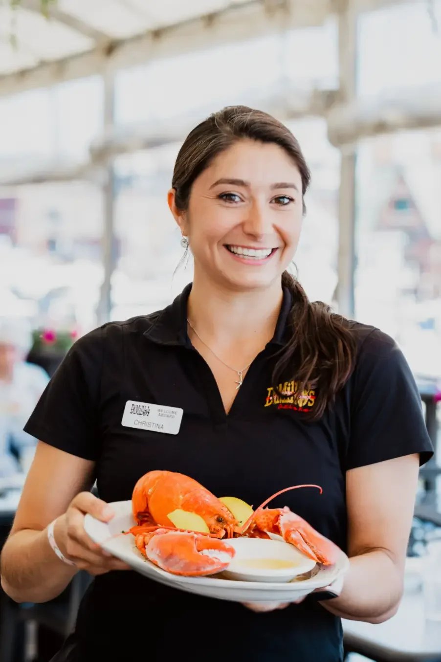 A woman in a black shirt holds a plate filled with cooked lobsters, smiling at the camera.
