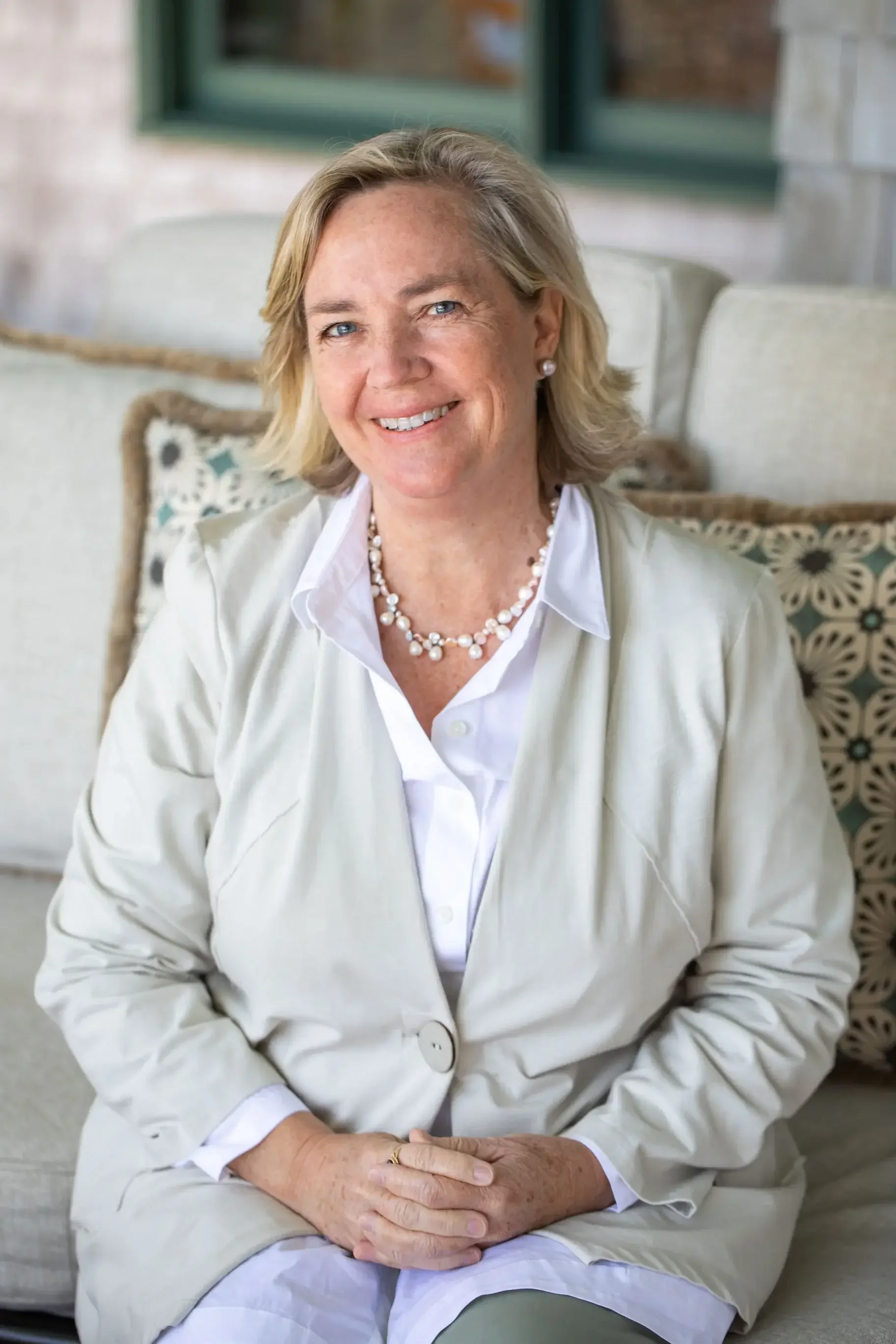 A woman wearing a white shirt and jacket sits comfortably on a couch, looking thoughtfully to the side