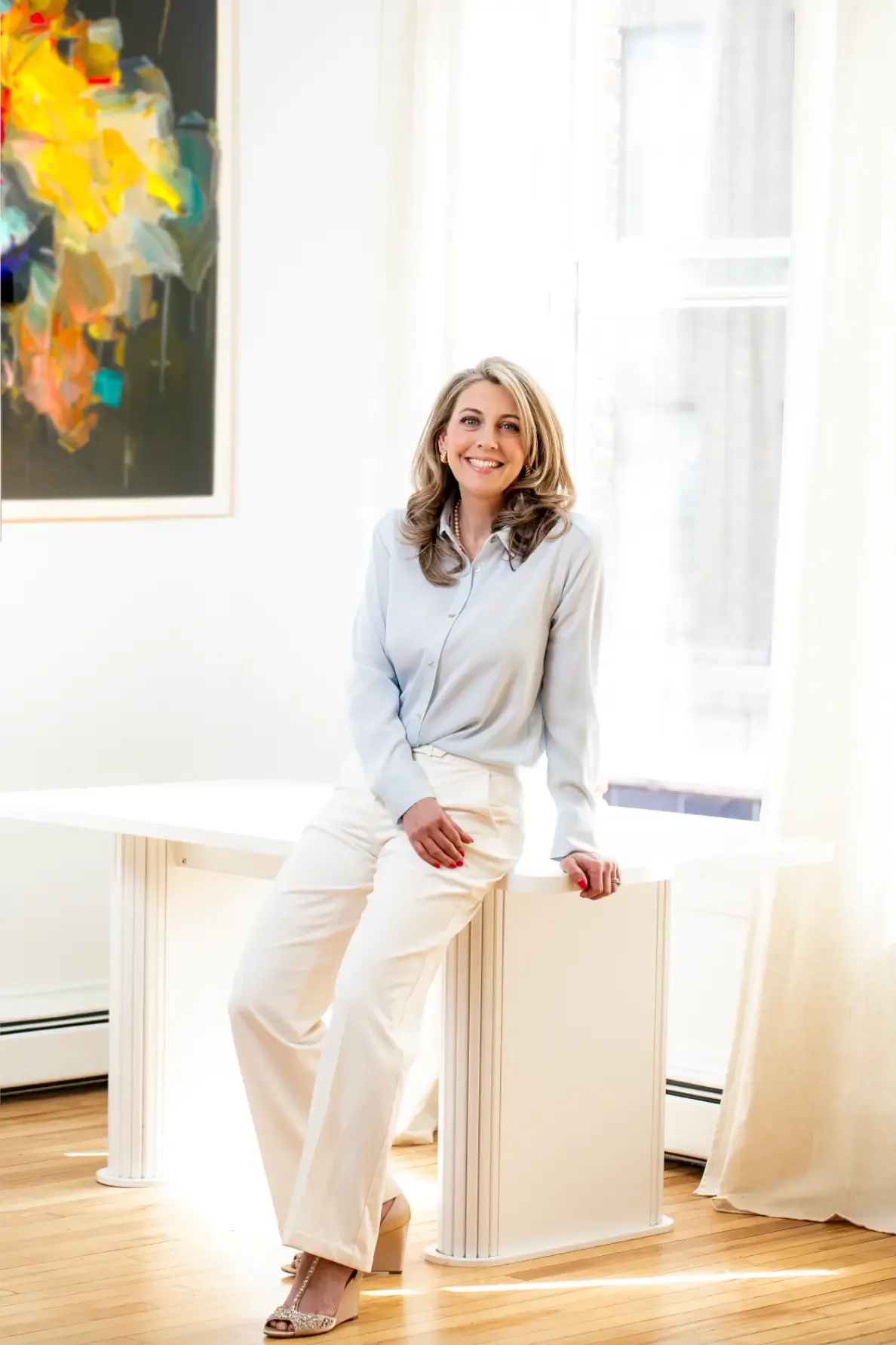 A woman wearing a blue shirt and white pants sits on a white table, smiling and looking towards the camera.