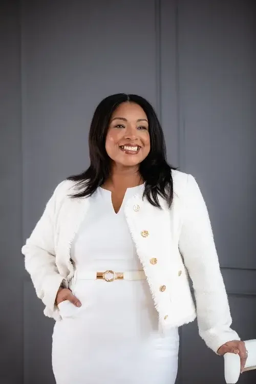 A woman in a white dress and jacket stands confidently in front of a chair, showcasing a stylish and elegant look.
