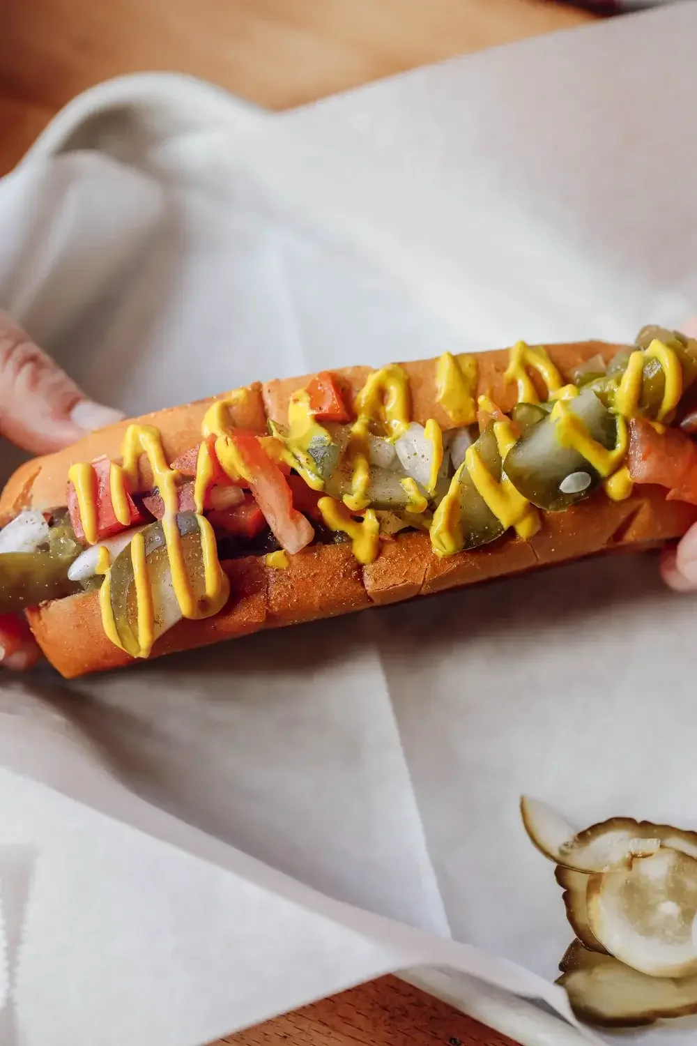 A hot dog topped with yellow mustard and sliced pickles on a white plate.
