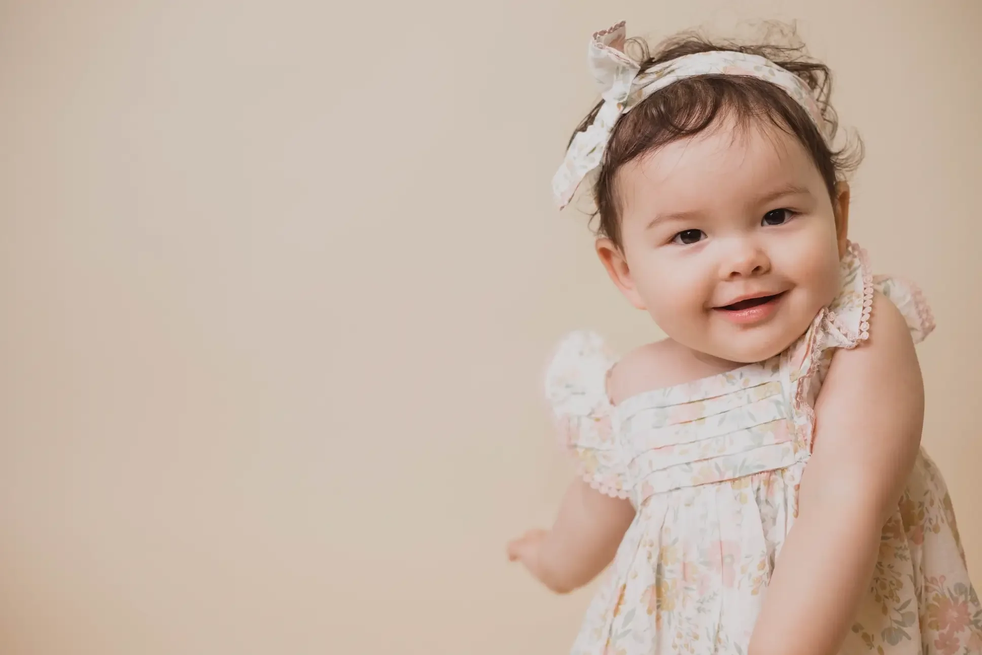 A baby girl wearing a floral dress smiles brightly at the camera, showcasing her joyful expression.