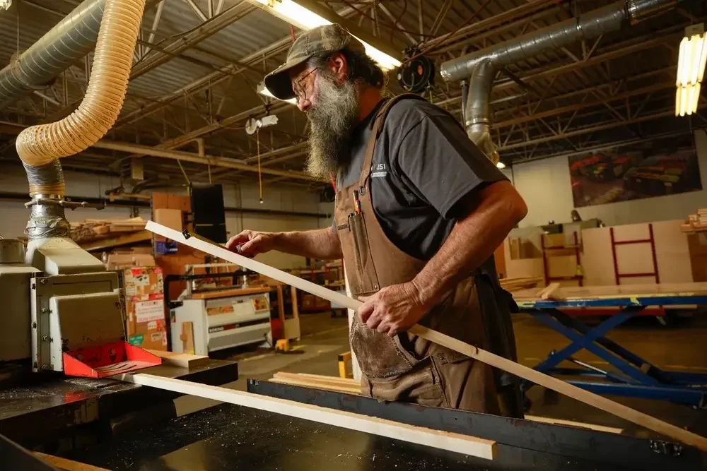 A bearded man wearing a hat focuses on a woodworking project, using tools and materials in a workshop setting.