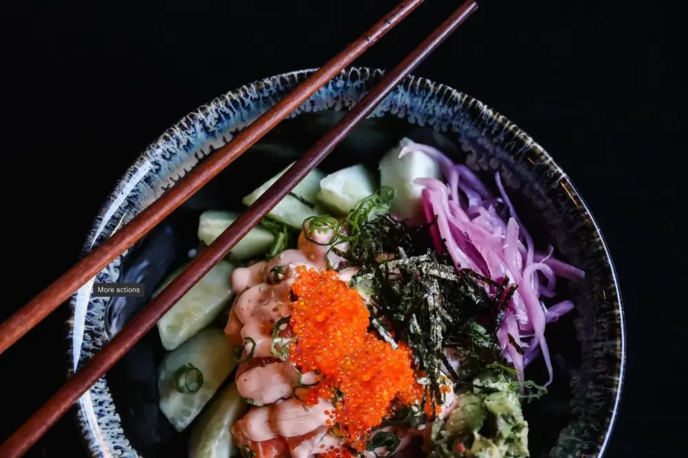A bowl of sushi featuring various rolls, accompanied by chopsticks and slices of cucumber.
