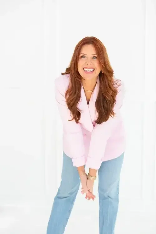 A woman wearing a pink jacket and jeans poses confidently for a photo, smiling at the camera.
