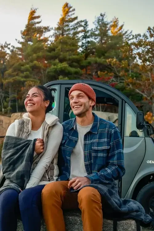 A couple sits on a bench beside a parked car, enjoying a moment together outdoors