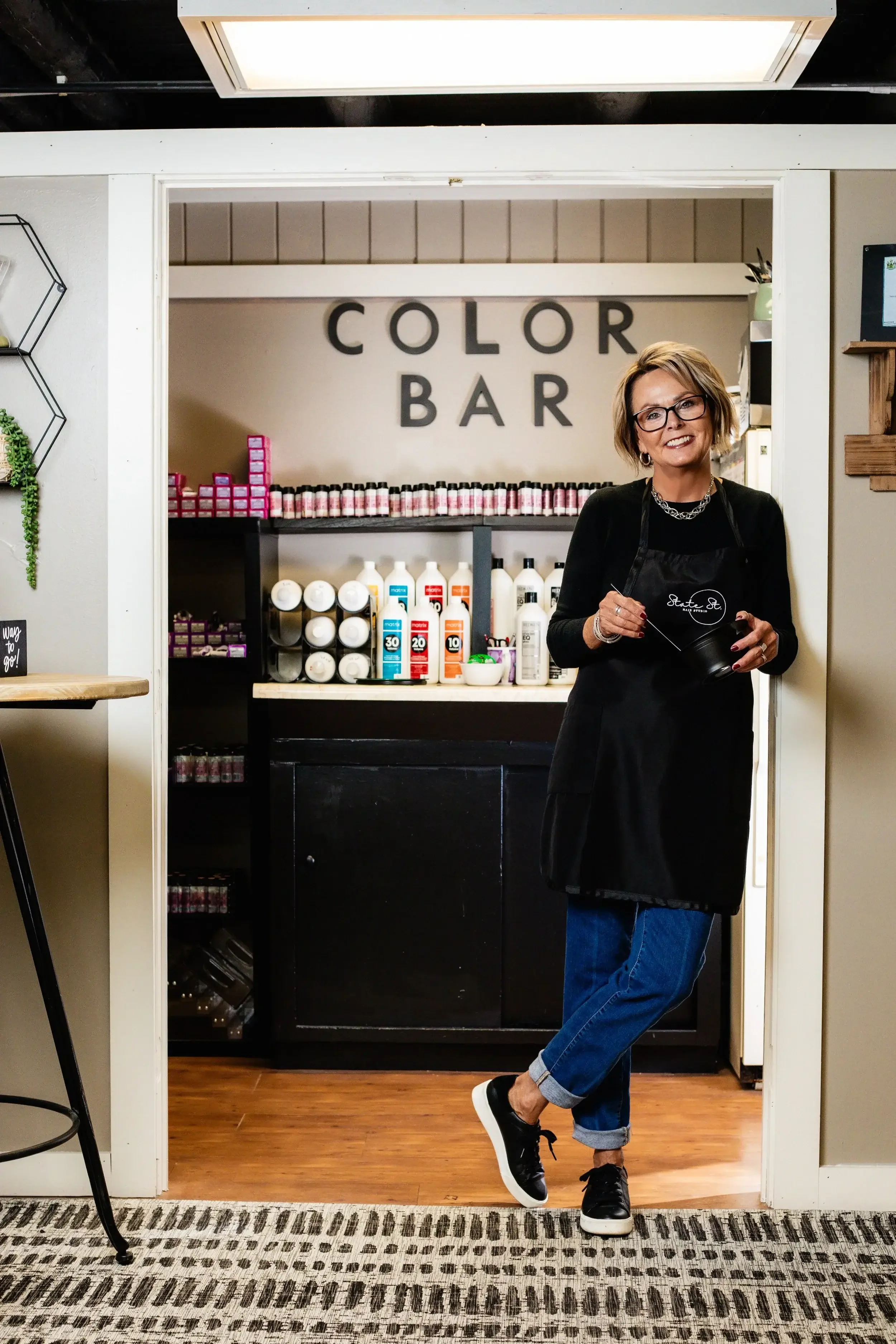 A woman stands in front of a door displaying a sign that reads "Color Bar," indicating the entrance to the establishment