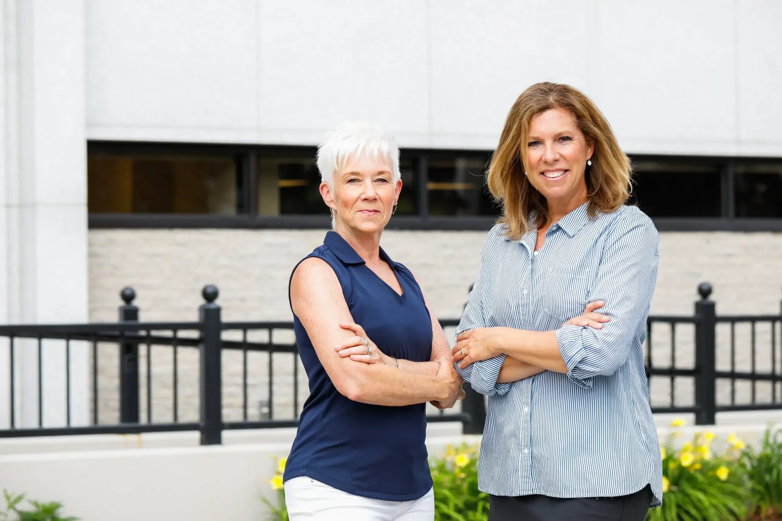 Two women stand together in front of a modern building, smiling and engaged in conversation.