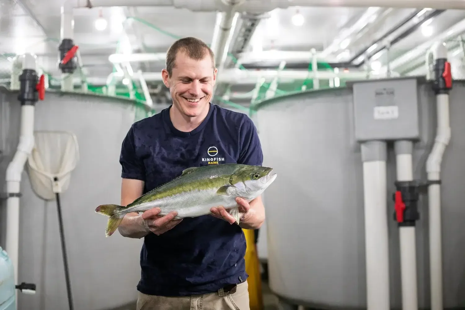 A man stands beside a fish tank, holding a fish with a smile, showcasing his catch.
