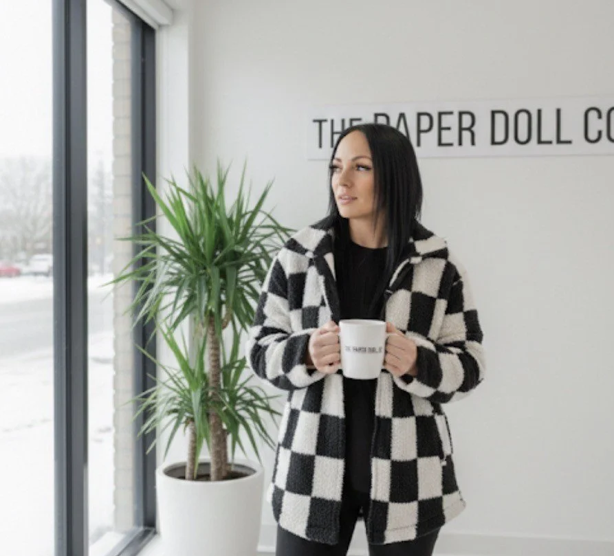 A woman with black hair in a checkered coat holding a white mug, standing indoors near a large window and a tall potted plant.