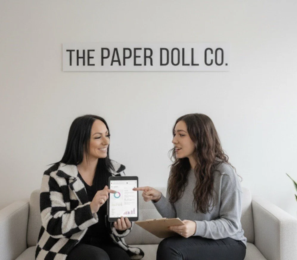 Two women sitting on a sofa in a modern office, discussing data displayed on a tablet. One woman is pointing at the tablet, and the other woman is holding a clipboard. A sign on the wall reads "The Paper Doll Co.".