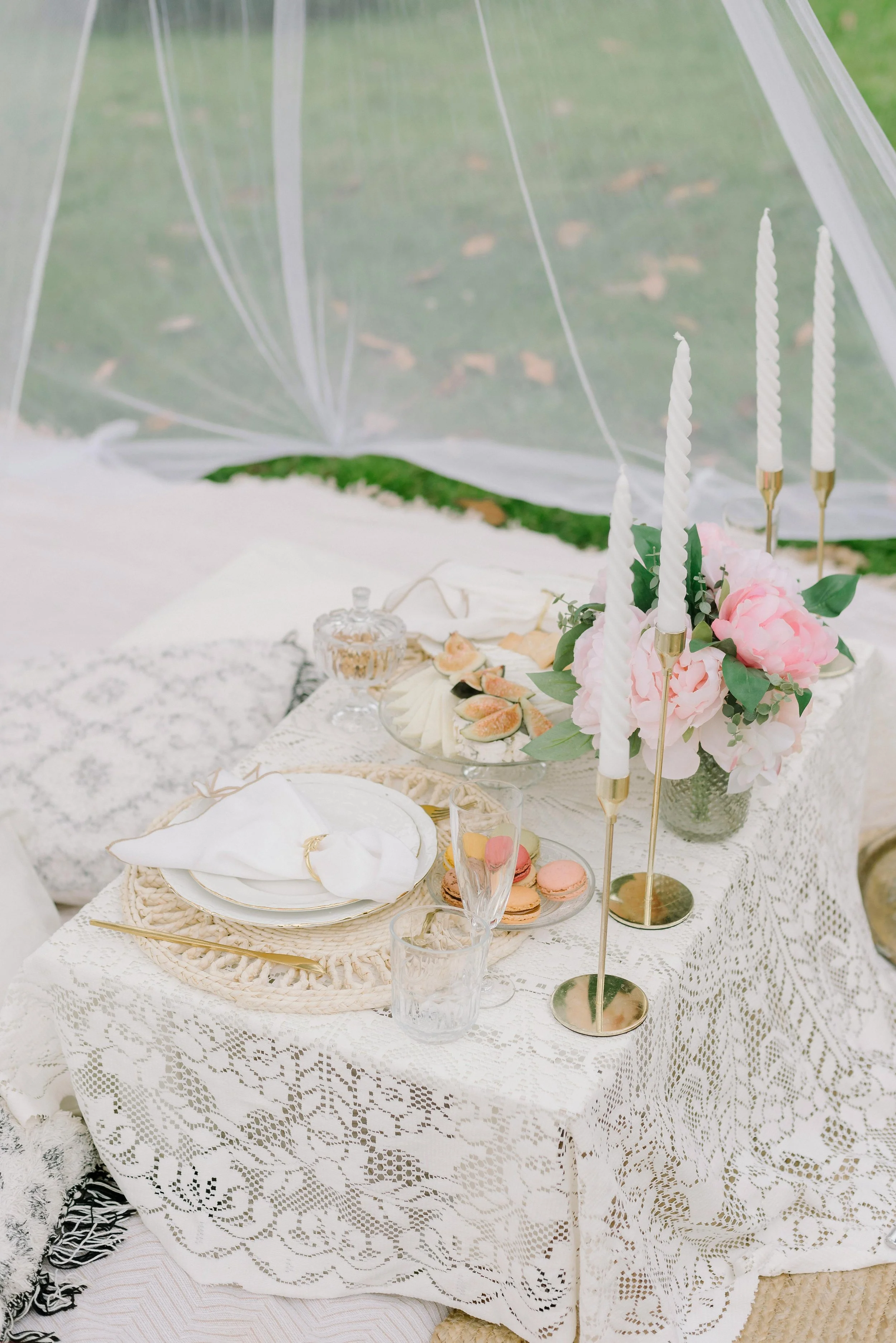 Outdoor picnic table with lace tablecloth, pink flowers, candles, macarons, cheese, figs, plates, and glasses.