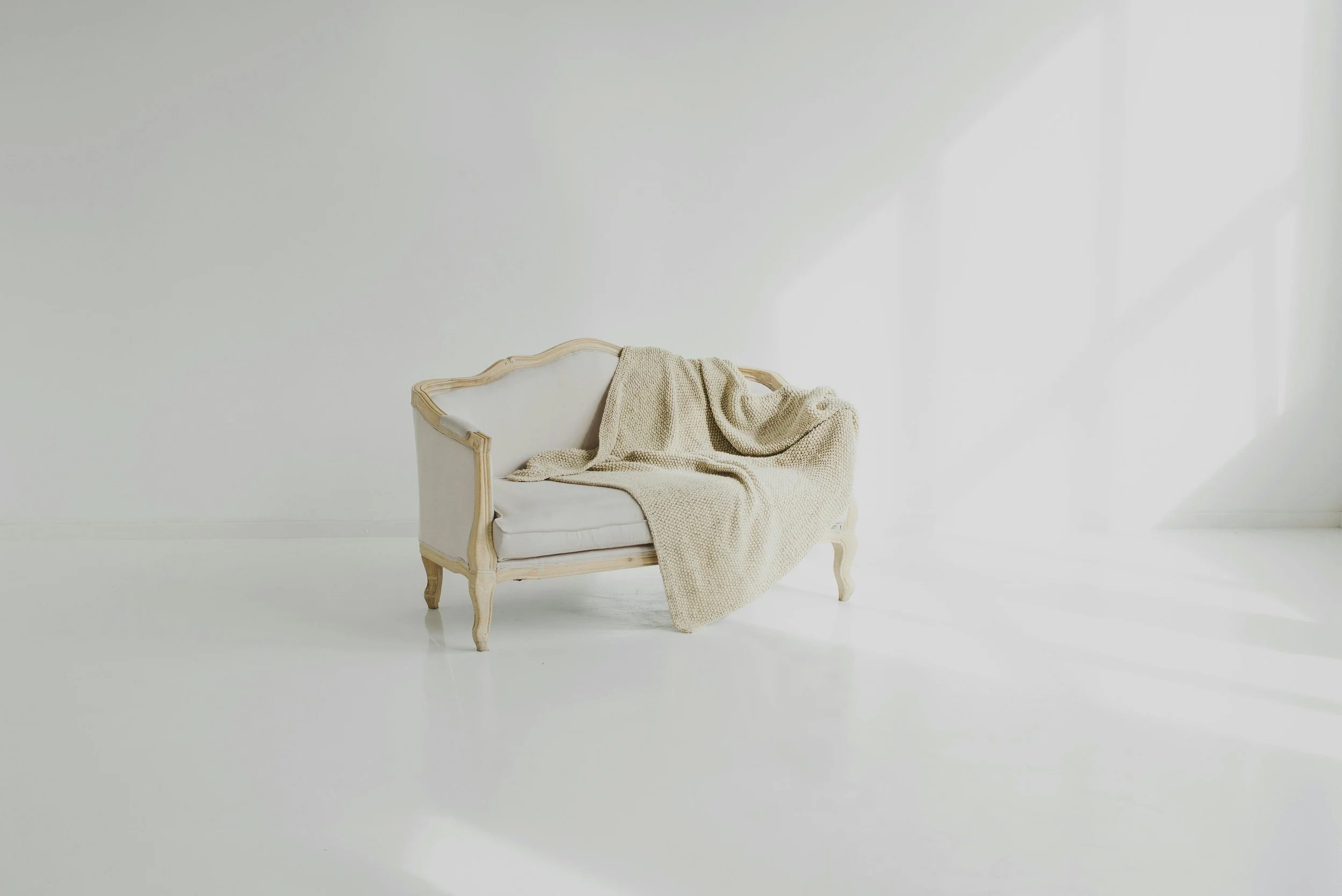A vintage-style white sofa with a wooden frame and carved legs, draped with a beige knitted blanket, set against a bright white wall in a minimalist room.