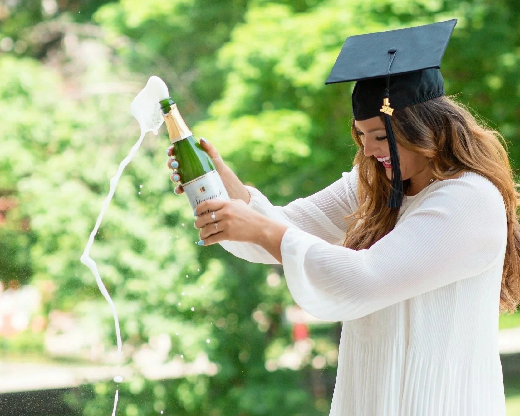 A woman wearing a graduation cap and gown celebrating outdoors by opening a bottle of champagne, with foam and bubbles spilling out.