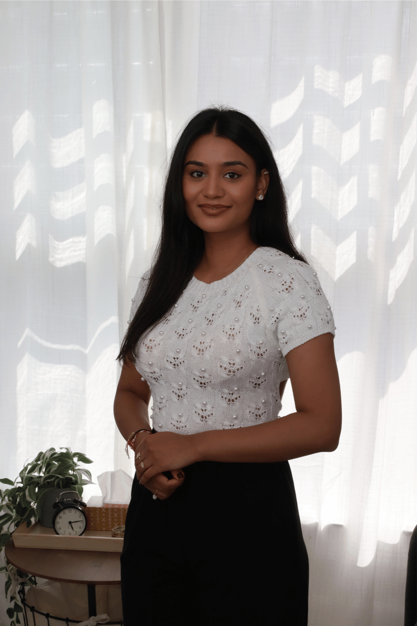 Psychotherapist standing in a softly lit office, wearing a white knit top and black skirt, with sheer curtains and a small plant in the background.