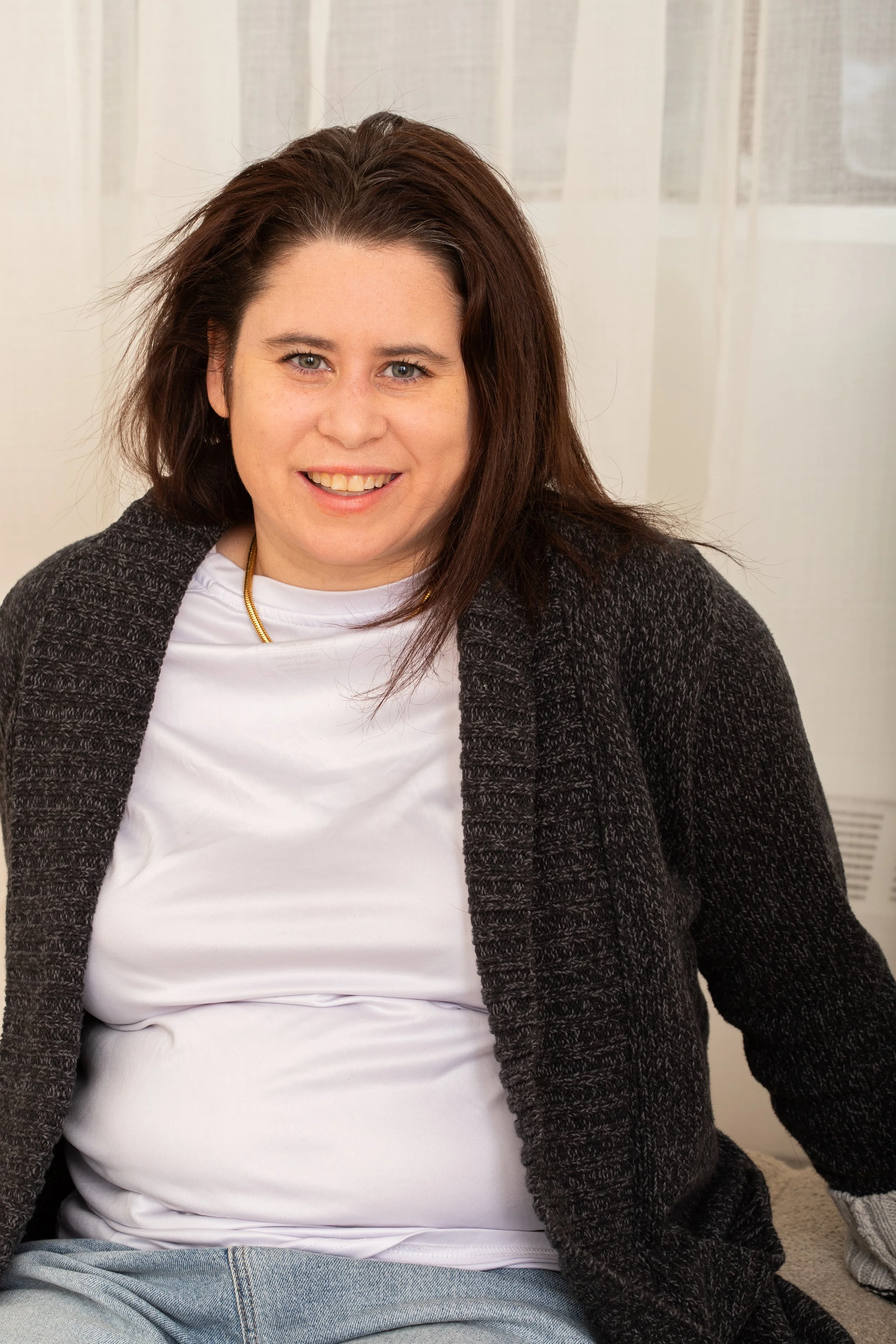 A woman with long brown hair, wearing a white shirt, a dark gray cardigan, and light blue jeans, sitting on a sofa and smiling at the camera.