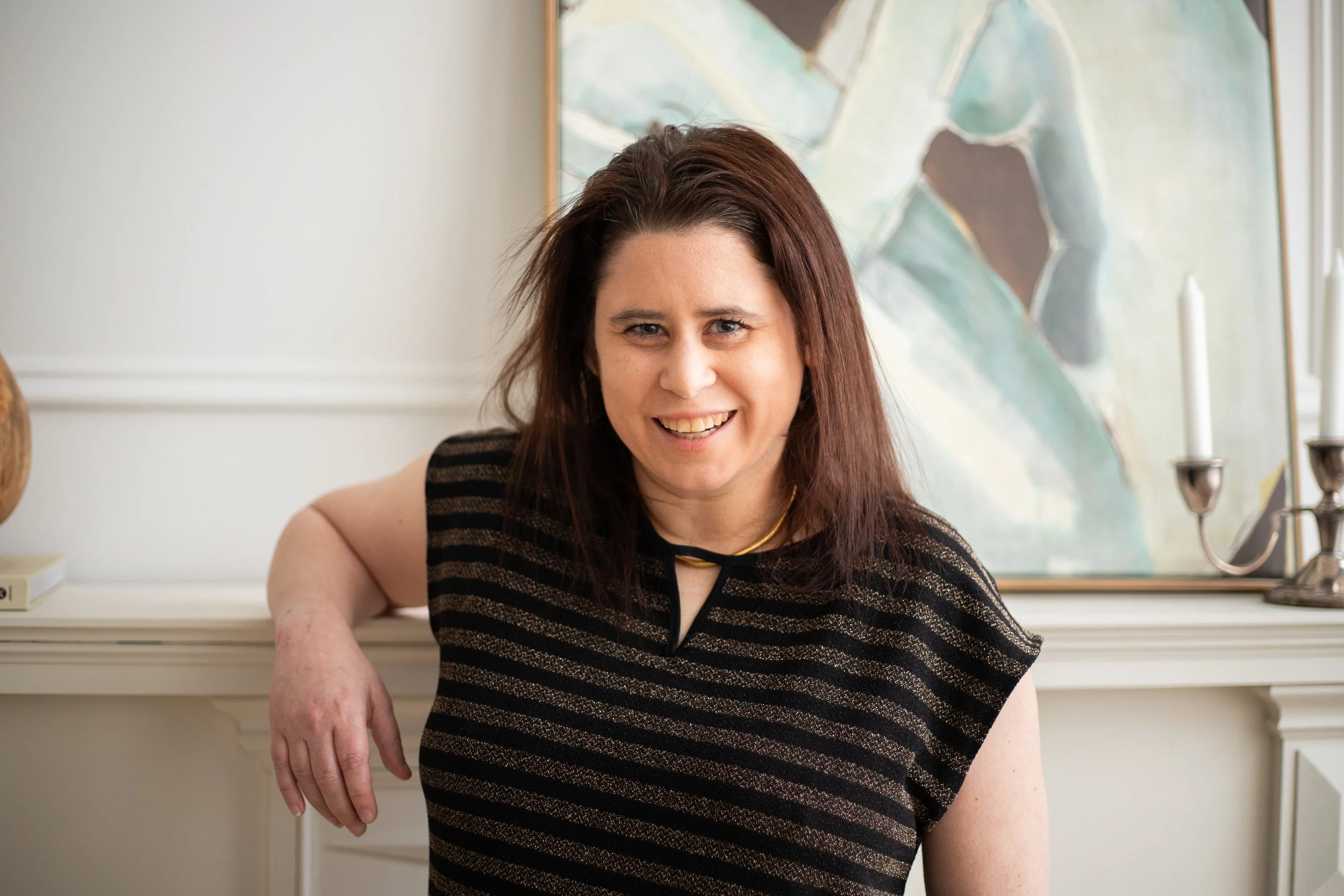 Smiling woman with dark red hair, wearing a black and gold striped top, sitting on a white fireplace mantel in front of an abstract painting and a candelabra.