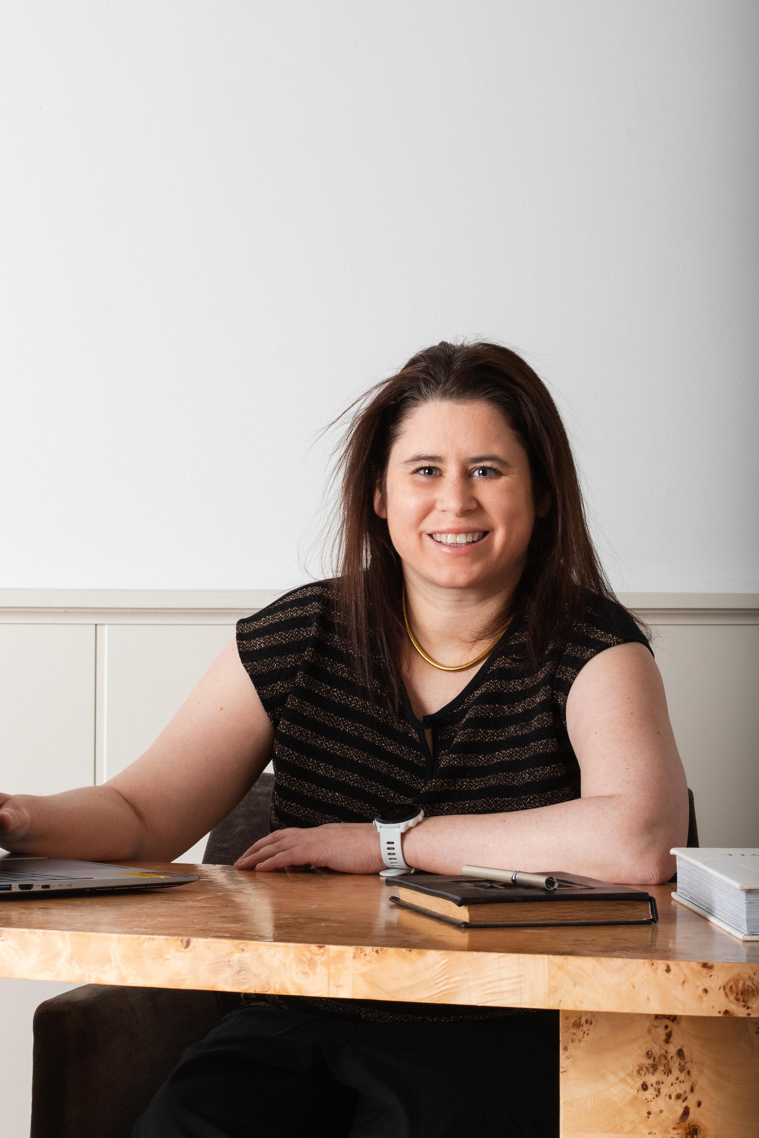 A woman sitting at a wooden desk smiling at the camera, with notebooks, a pen, and a laptop on the desk.