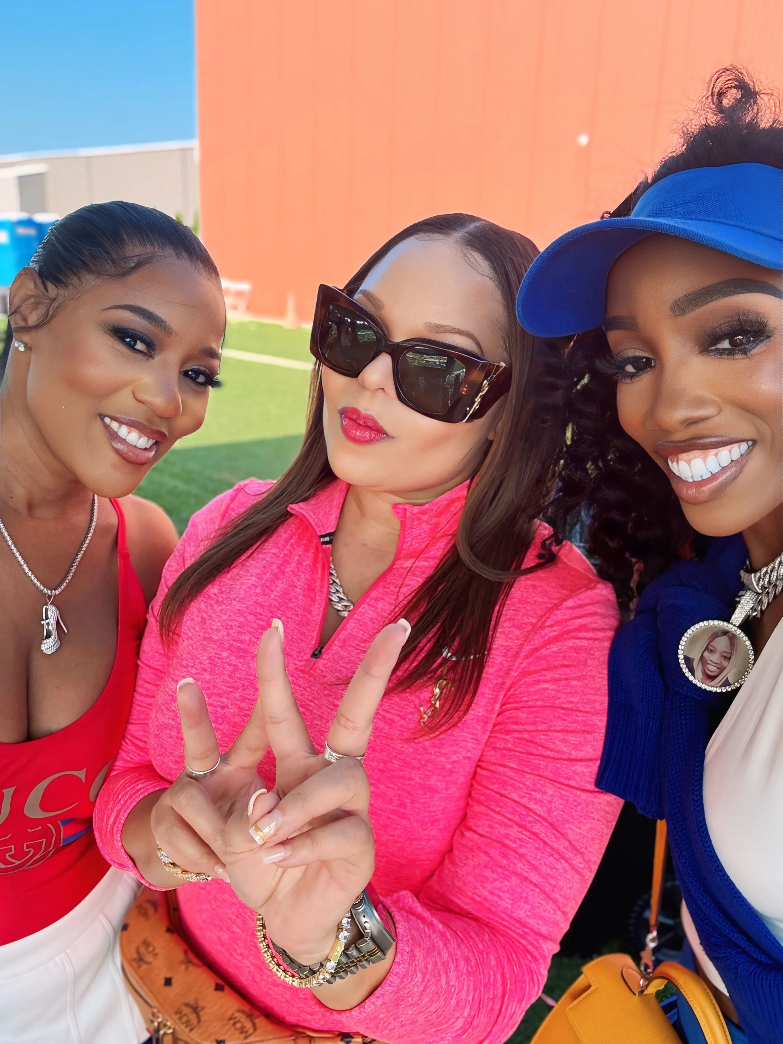 Three women taking a selfie together outdoors with a background of a red wall and blue sky. The woman in the middle is wearing large sunglasses and a pink jacket, making a peace sign with her fingers. The woman on the left has her hair slicked back a
