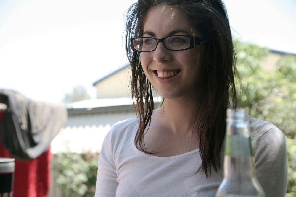 Young woman with glasses smiling outdoors with greenery and a building in the background.